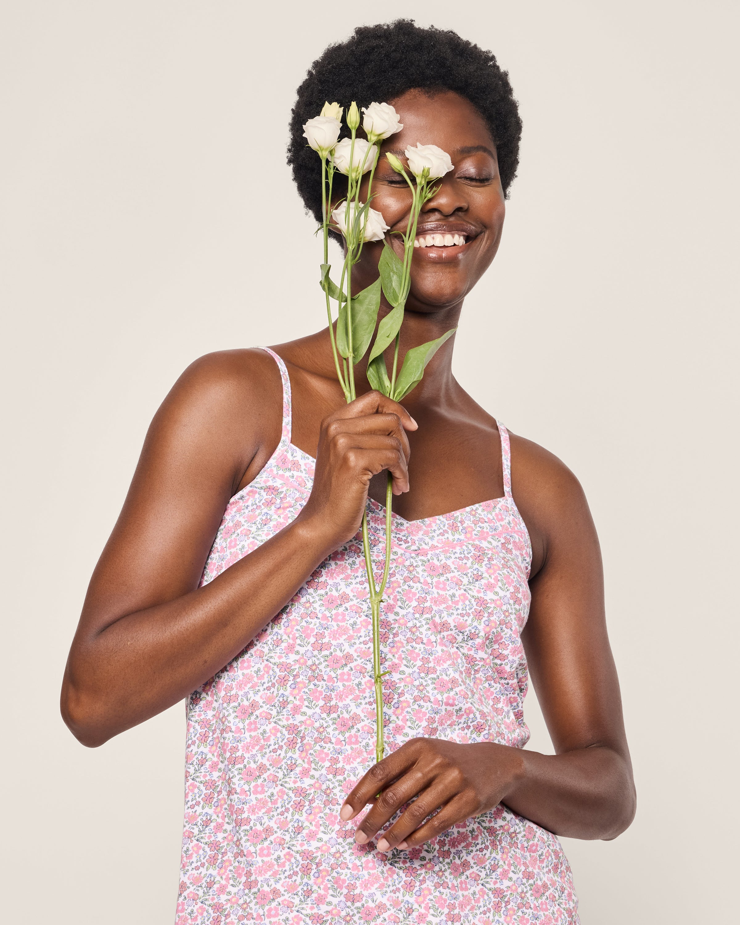 A woman wearing the Womens Pima Sylvie Short Set in Fleurs de Rose by Petite Plume smiles and holds white flowers near her face against a light background.