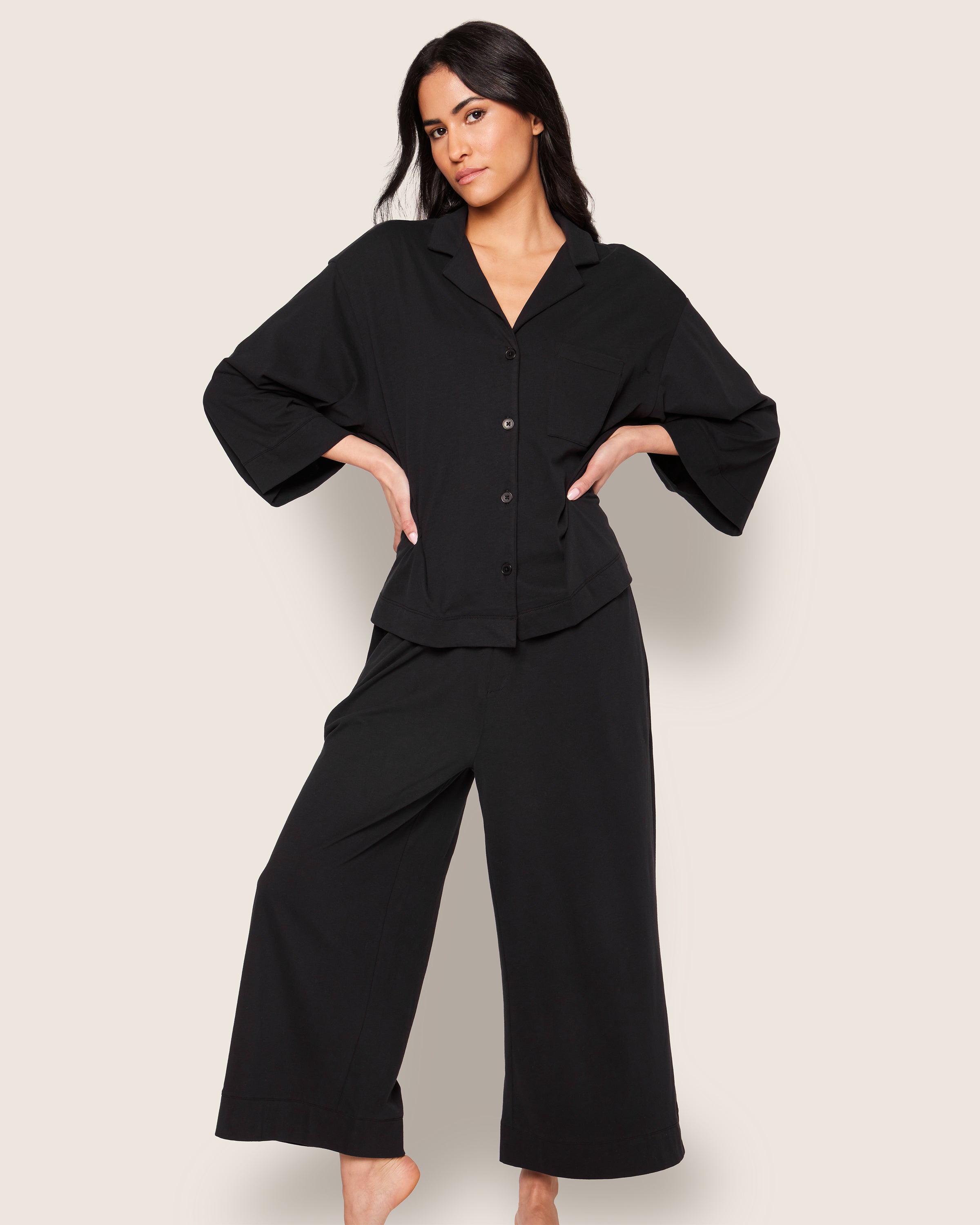 Modeling relaxed elegance, a person poses barefoot against an off-white background in The Olivia Wide Leg Pima Pajama Set from Petite Plume, featuring a loose-fitting black button-up top and wide-legged pants crafted from luxurious Peruvian Pima cotton.