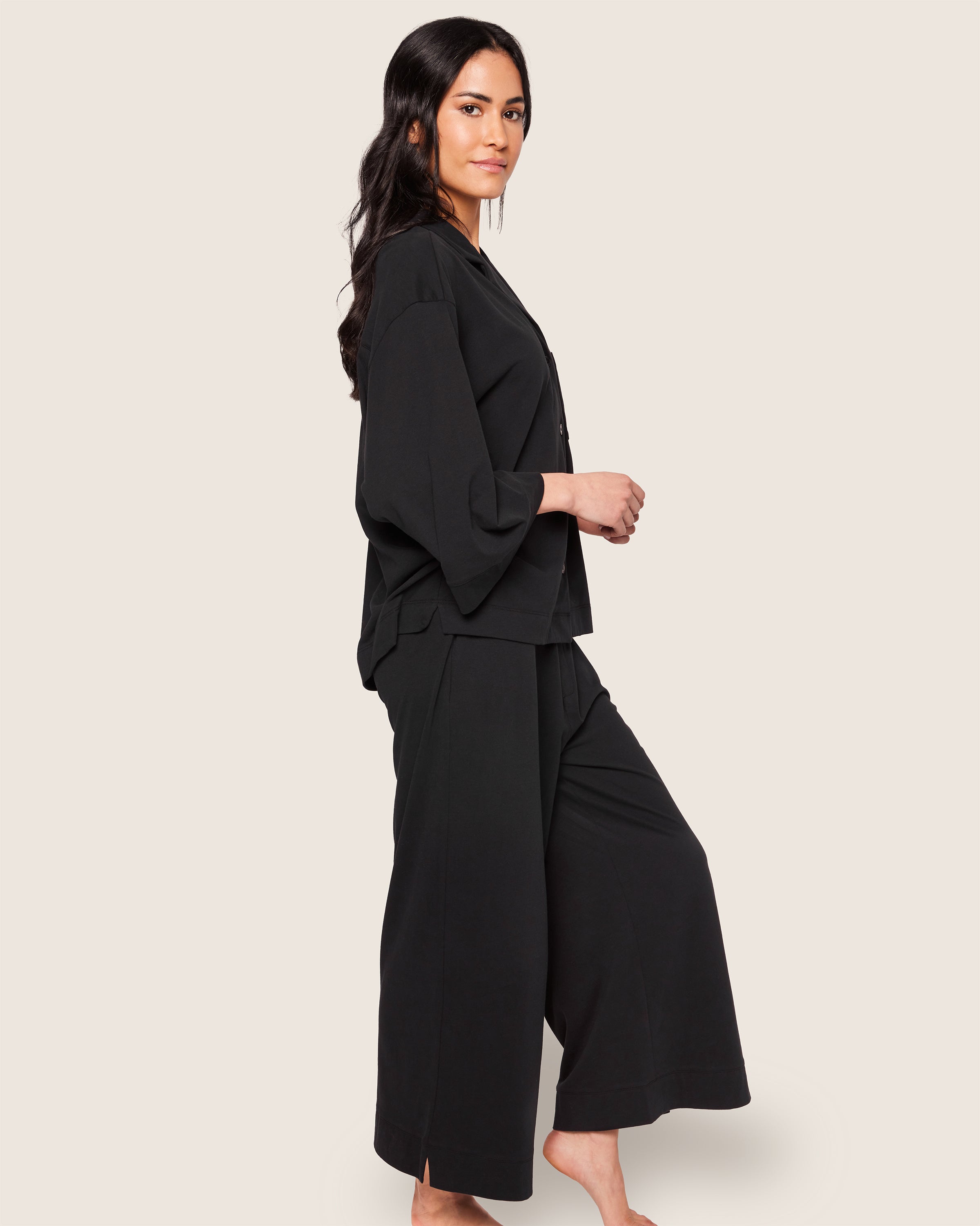 A woman stands in a side pose against a plain background wearing The Olivia Wide Leg Pima Pajama Set in Black by Petite Plume, showcasing the luxurious comfort of its long-sleeved top and wide-leg pants. Her long dark hair cascades as she looks forward.