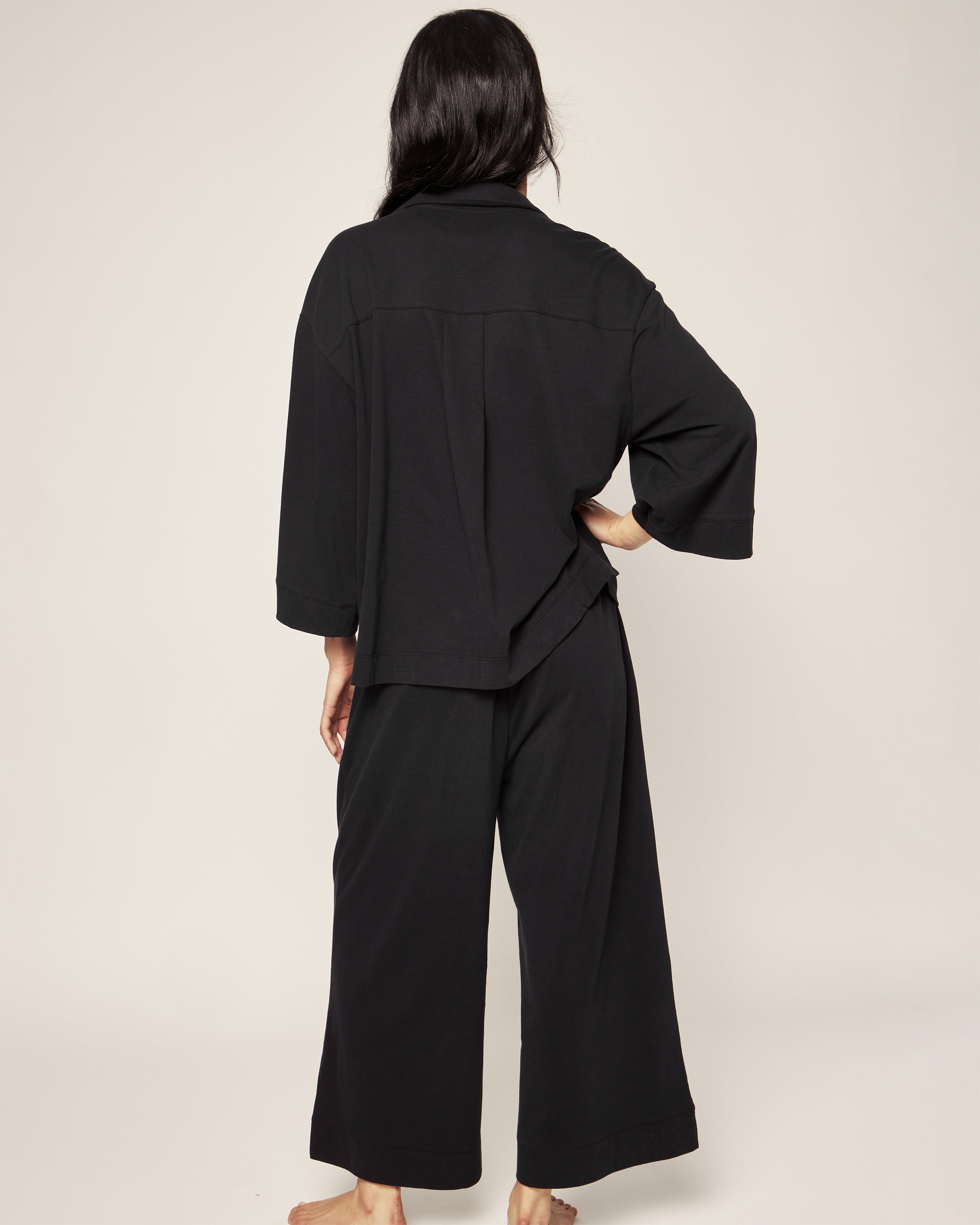 A person with long dark hair, facing away, is wrapped in The Olivia Wide Leg Pima Pajama Set in Black by Petite Plume. This luxurious sleepwear set features a loose jersey knit design with wide-leg pants and a long-sleeved top, exuding effortless elegance against a neutral background.