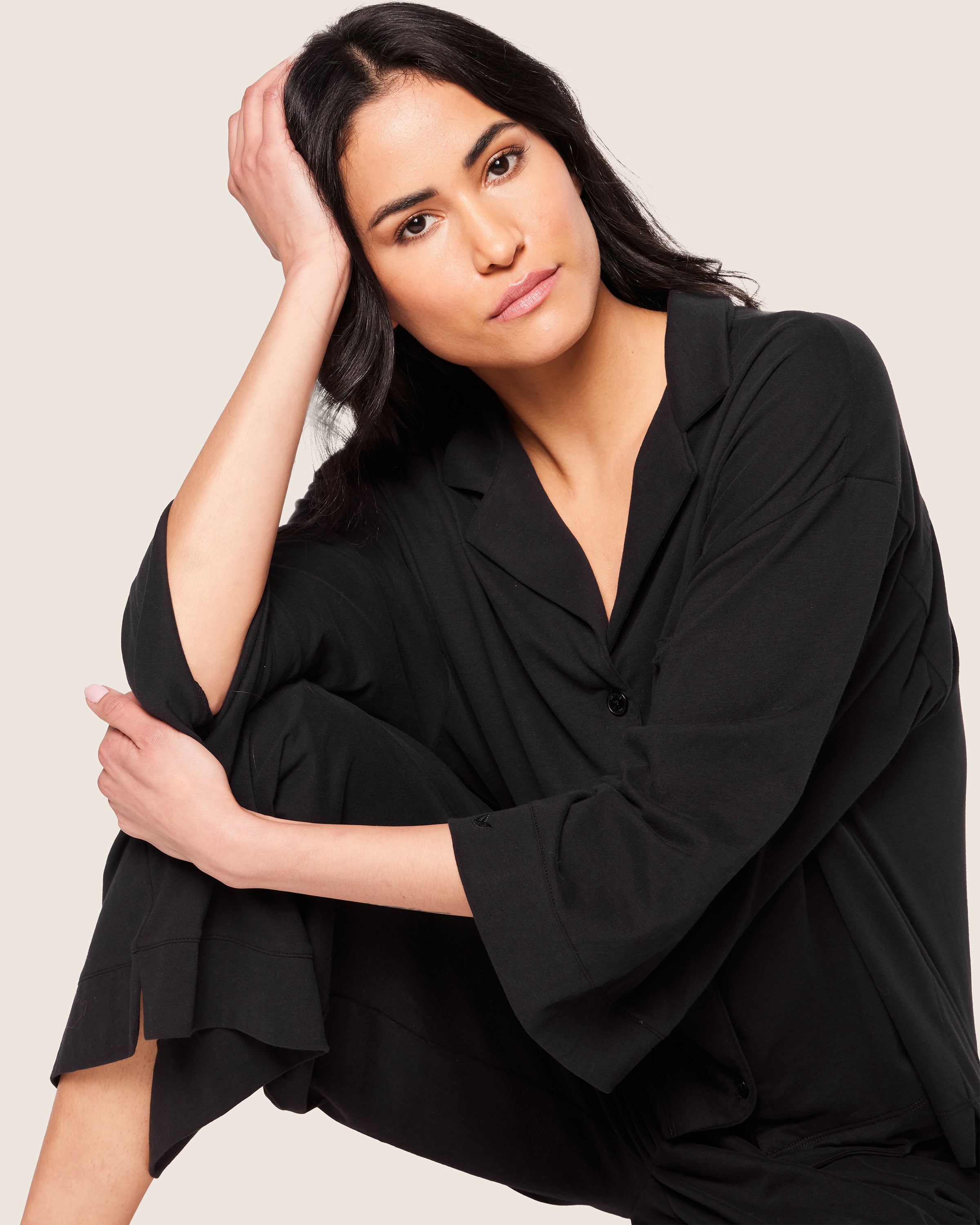 A woman with long dark hair sits, wearing the Petite Plume Olivia Wide Leg Pima Pajama Set in black. She rests her head on her hand, looking directly at the camera with a neutral expression, against a light and plain background.