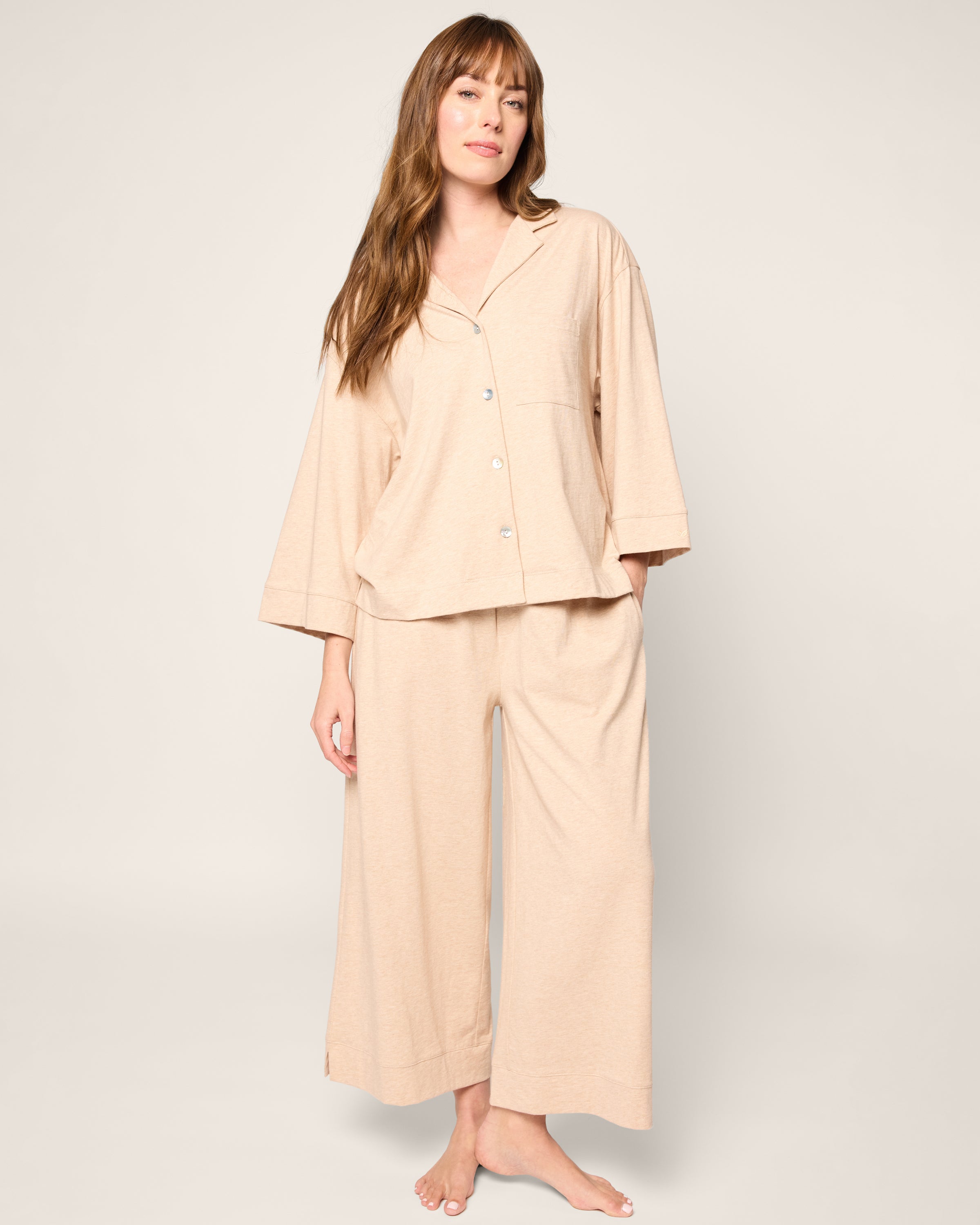 A person in The Olivia Wide Leg Pima Pajama Set in Camel by Petite Plume poses against a simple, light-colored backdrop. They have long hair and are barefoot, conveying a relaxed and casual expression.