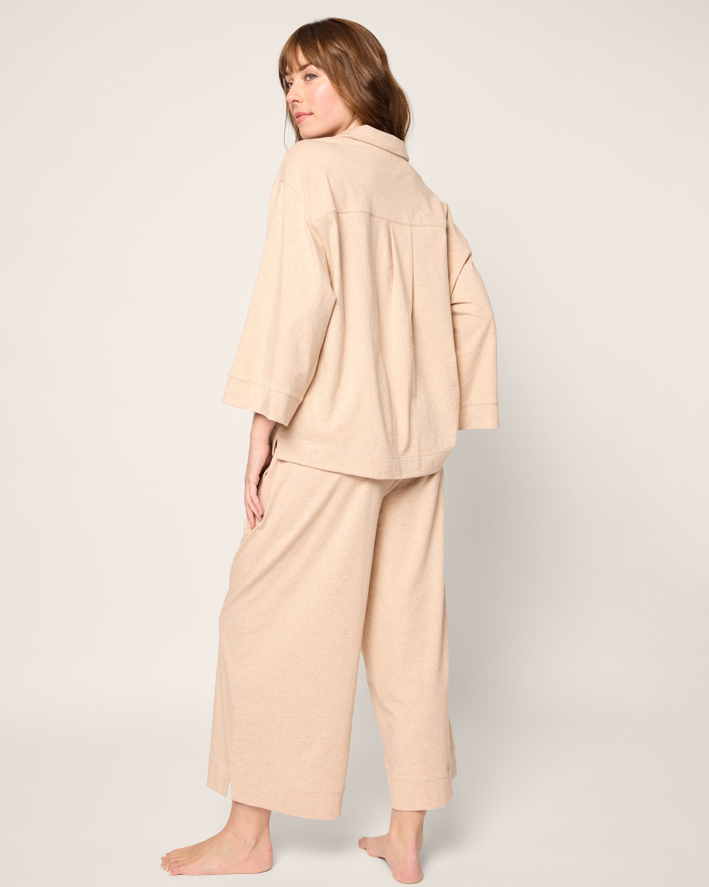 A person barefoot, wearing the Olivia Wide Leg Pima Pajama Set in Camel by Petite Plume, stands against a plain background. They are turned slightly to the side, looking over their shoulder.