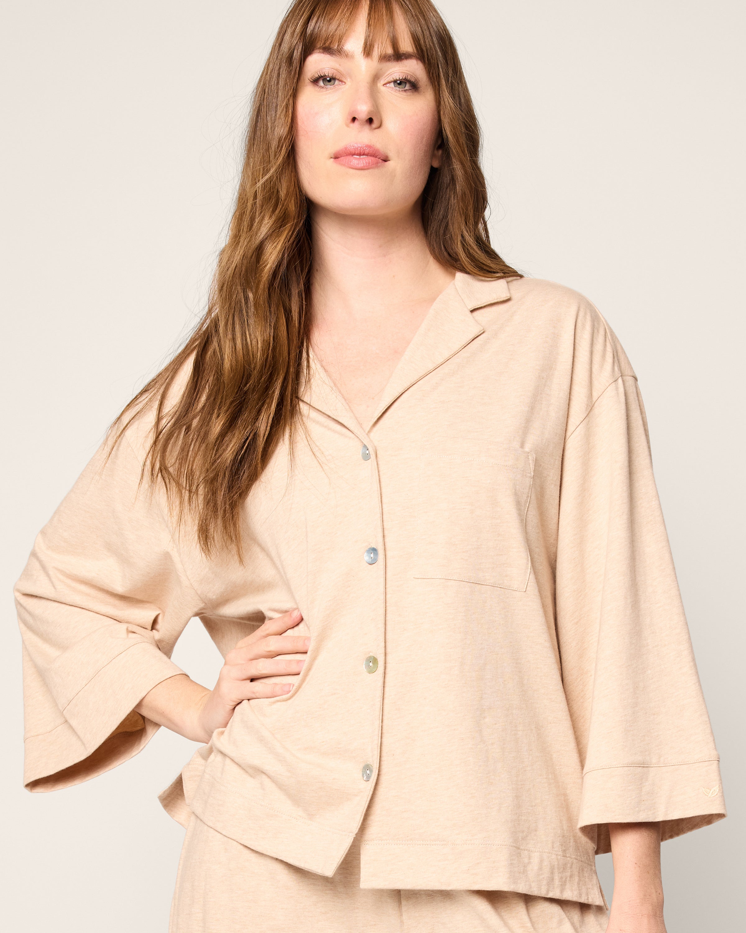 A woman with long brown hair wears The Olivia Wide Leg Pima Pajama Set in Camel by Petite Plume, featuring a loose-fitting top made from soft Peruvian Pima cotton. She stands confidently against a plain background, looking directly at the camera with one hand on her hip.