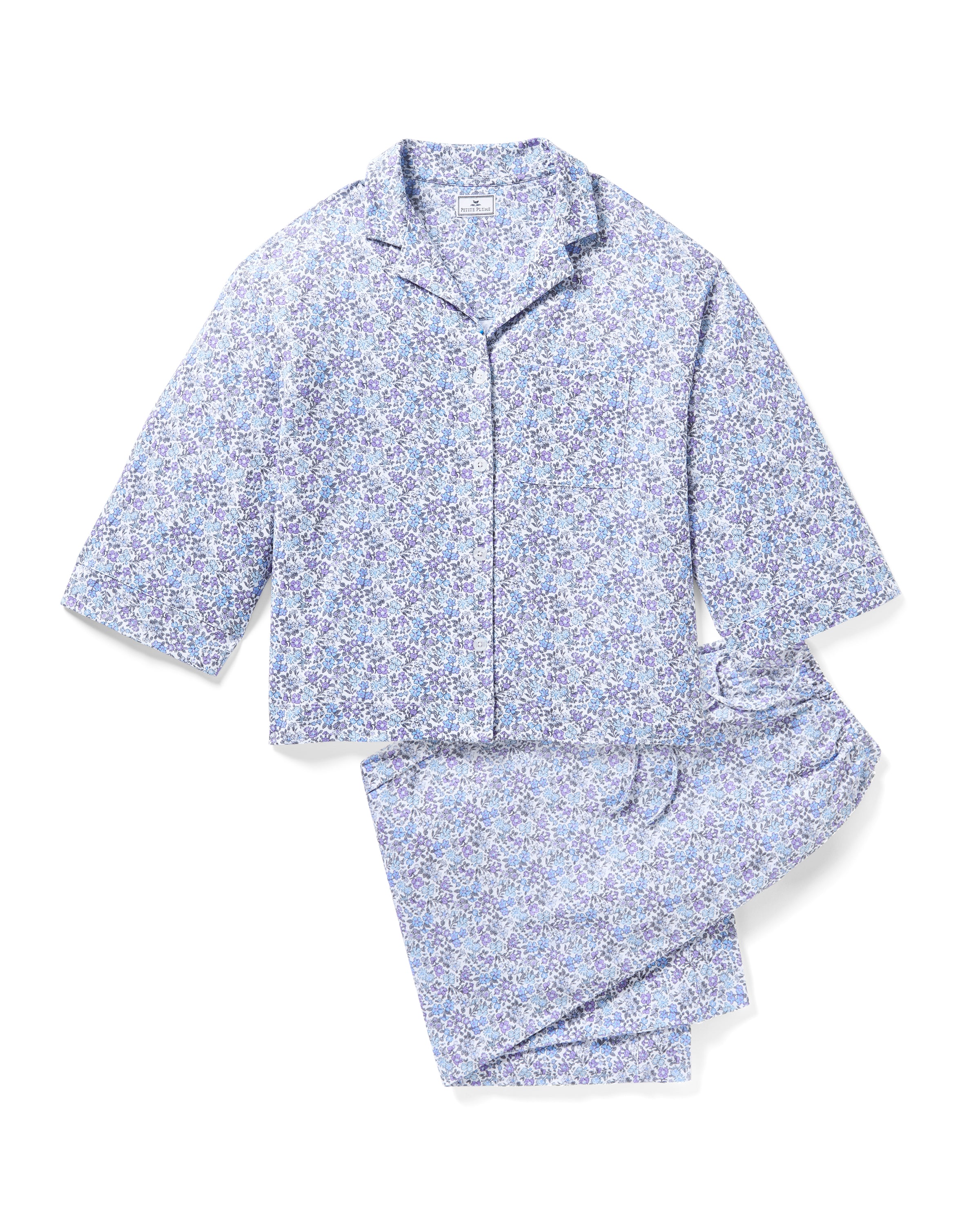 The Olivia Wide Leg Pima Pajama Set in Fleur dAzur by Petite Plume is a blue floral-patterned set, made from luxurious Peruvian Pima cotton. It includes a button-up top with a collar and long sleeves, and matching wide-leg pants, all displayed flat on a white background.