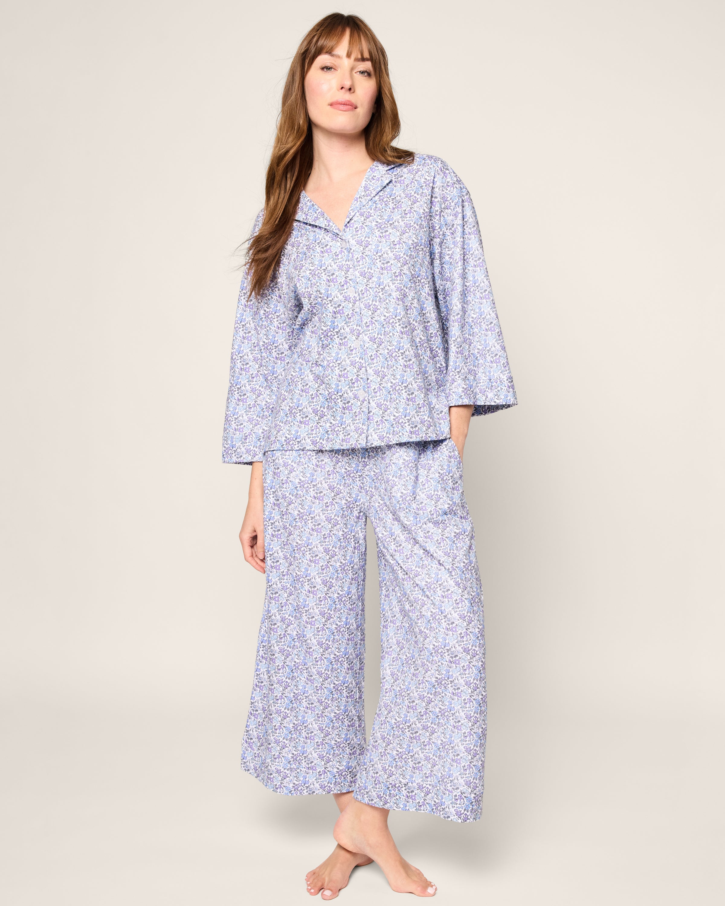 A person stands barefoot in Petite Plumes Olivia Wide Leg Pima Pajama Set in Fleur dAzur, featuring a small floral pattern with a long-sleeve button-up top and comfy wide-leg pants against a plain, light background.