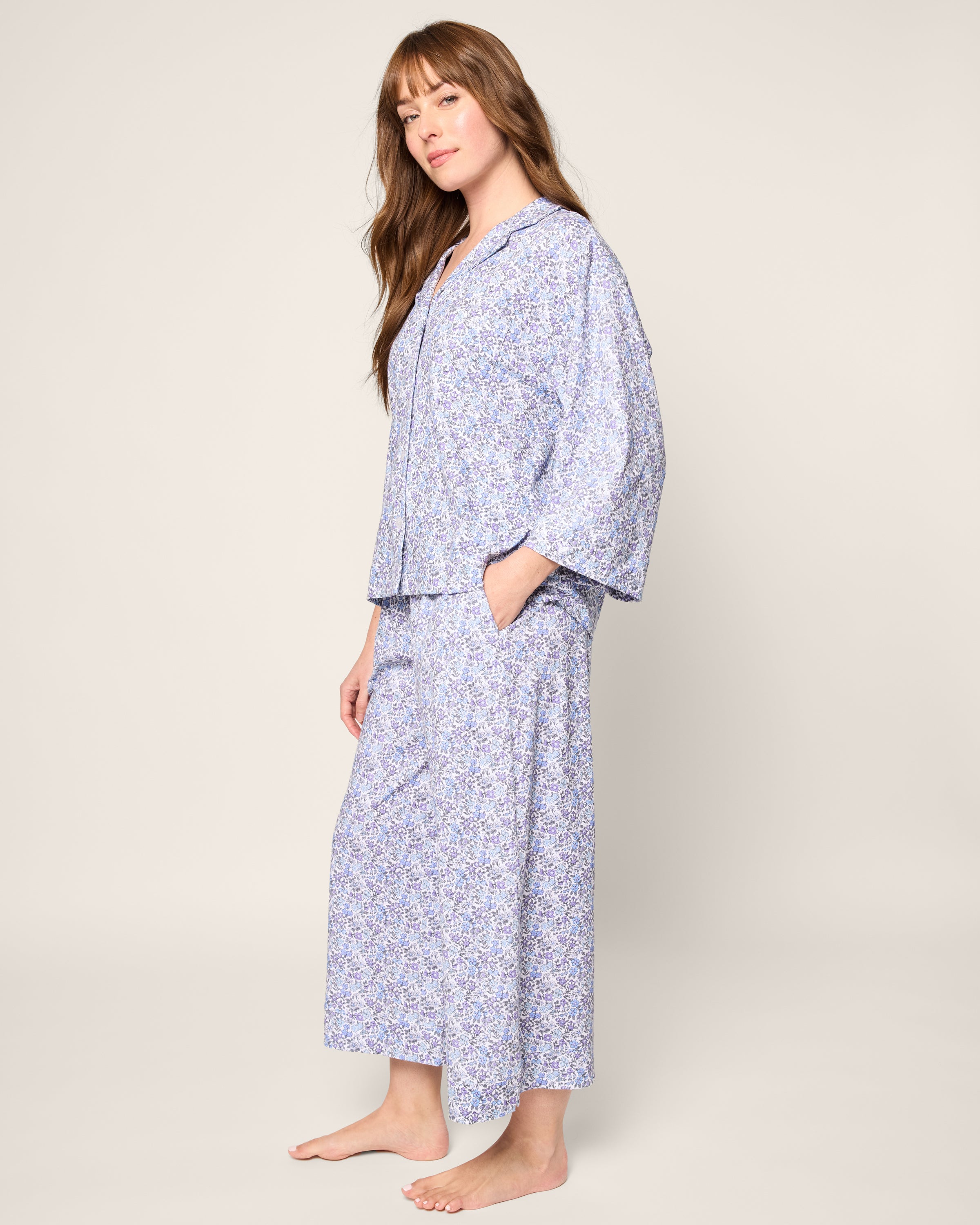 A person with long hair stands barefoot, wearing Petite Plumes The Olivia Wide Leg Pima Pajama Set in Fleur dAzur, made from soft Peruvian Pima cotton. They face slightly sideways with hands in pockets against a plain background.