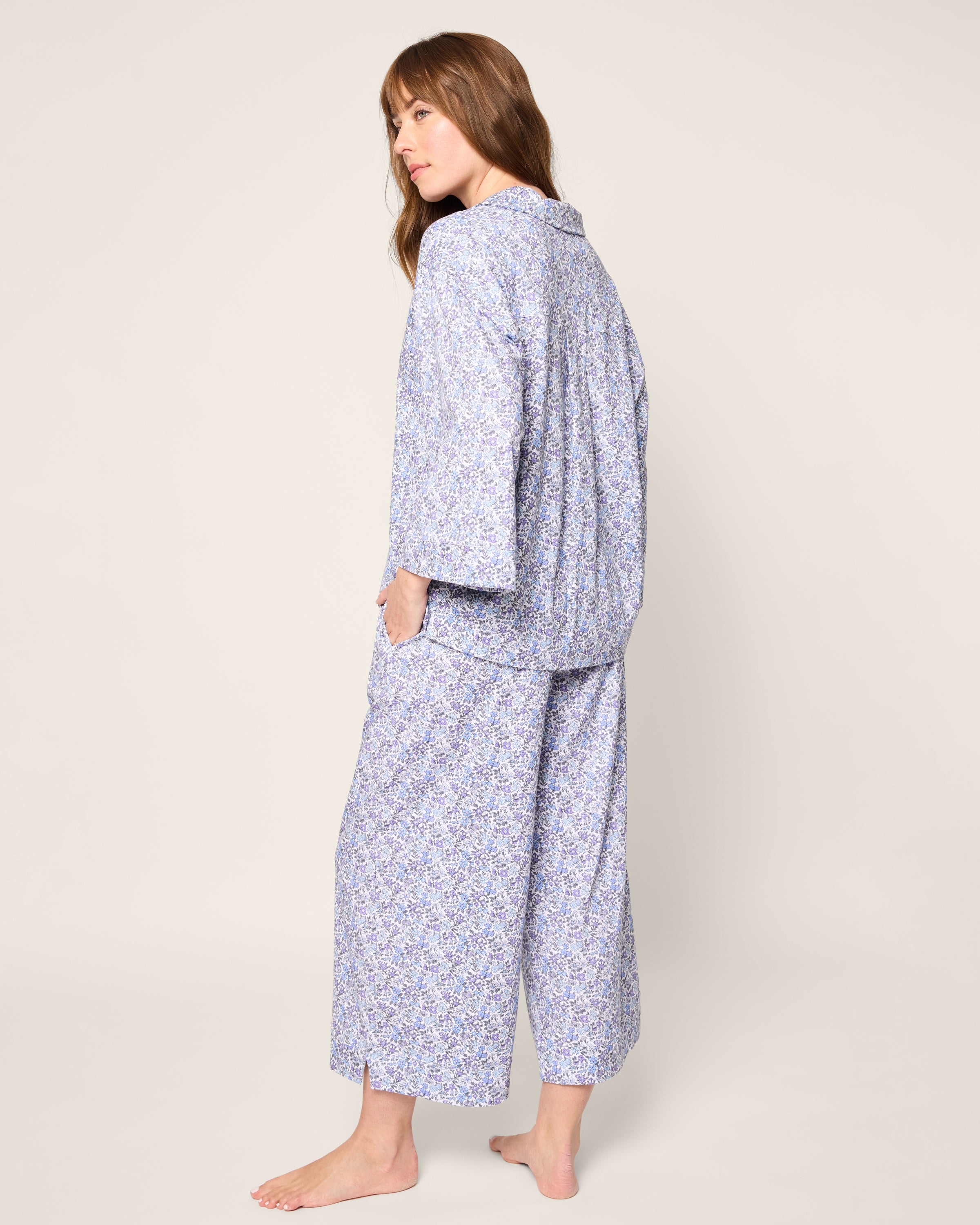 A person with long hair stands barefoot, slightly turned away, wearing Petite Plumes Olivia Wide Leg Pima Pajama Set in Fleur dAzur. The loose pajama set is crafted from soft Peruvian Pima cotton, featuring wide sleeves and wide-leg pants against a plain, light background.