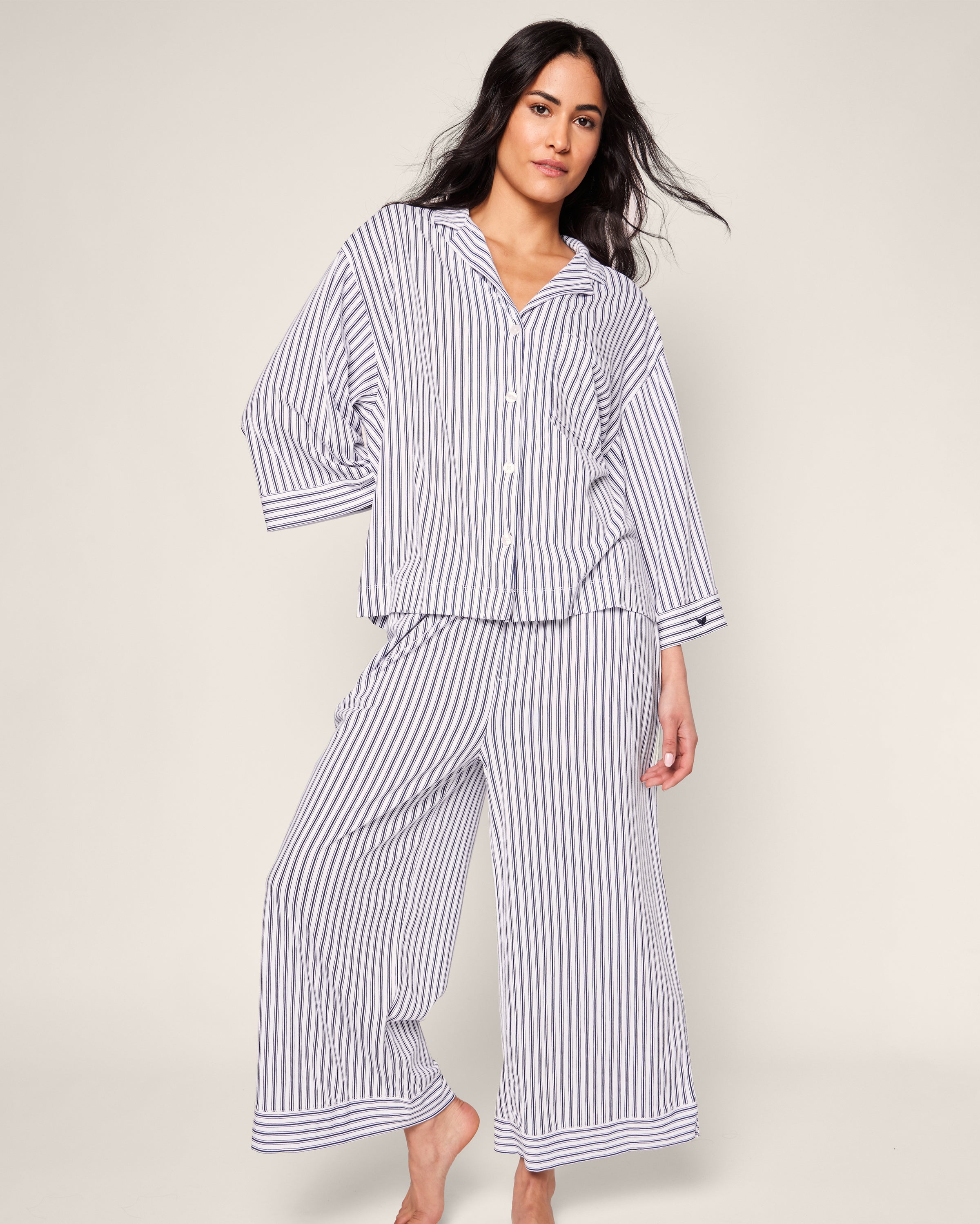 A person with long dark hair is wearing Petite Plumes Olivia Wide Leg Pima Pajama Set in Navy French Ticking, crafted from luxurious Peruvian Pima cotton. The button-up shirt and wide-legged pants have vertical stripes in alternating dark and light colors. They stand barefoot against a plain background.