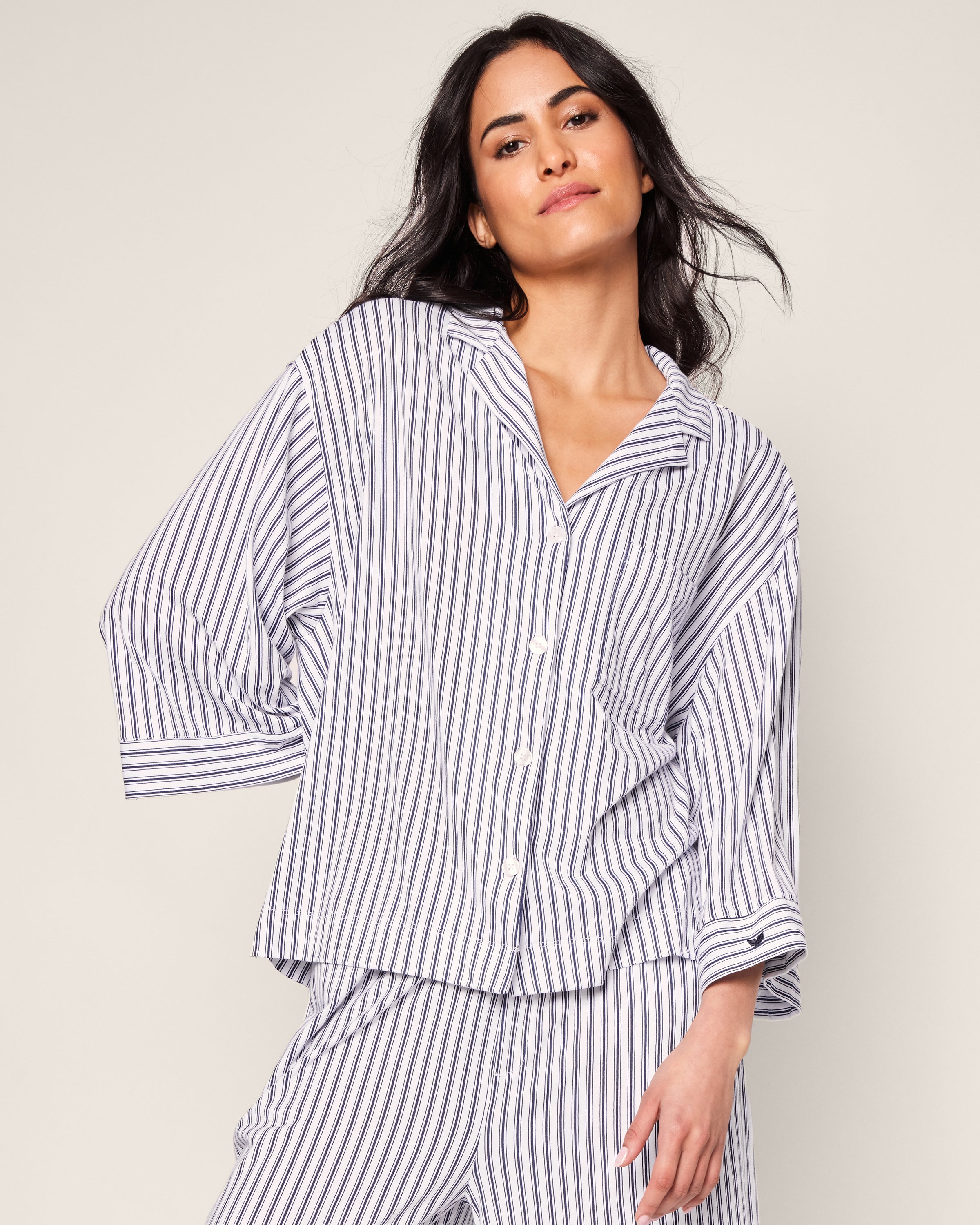 A person is wearing the Petite Plumes Olivia Wide Leg Pima Pajama Set in Navy French Ticking. They stand against a plain background, head slightly tilted, hands on hips, showcasing its relaxed and comfortable fit.