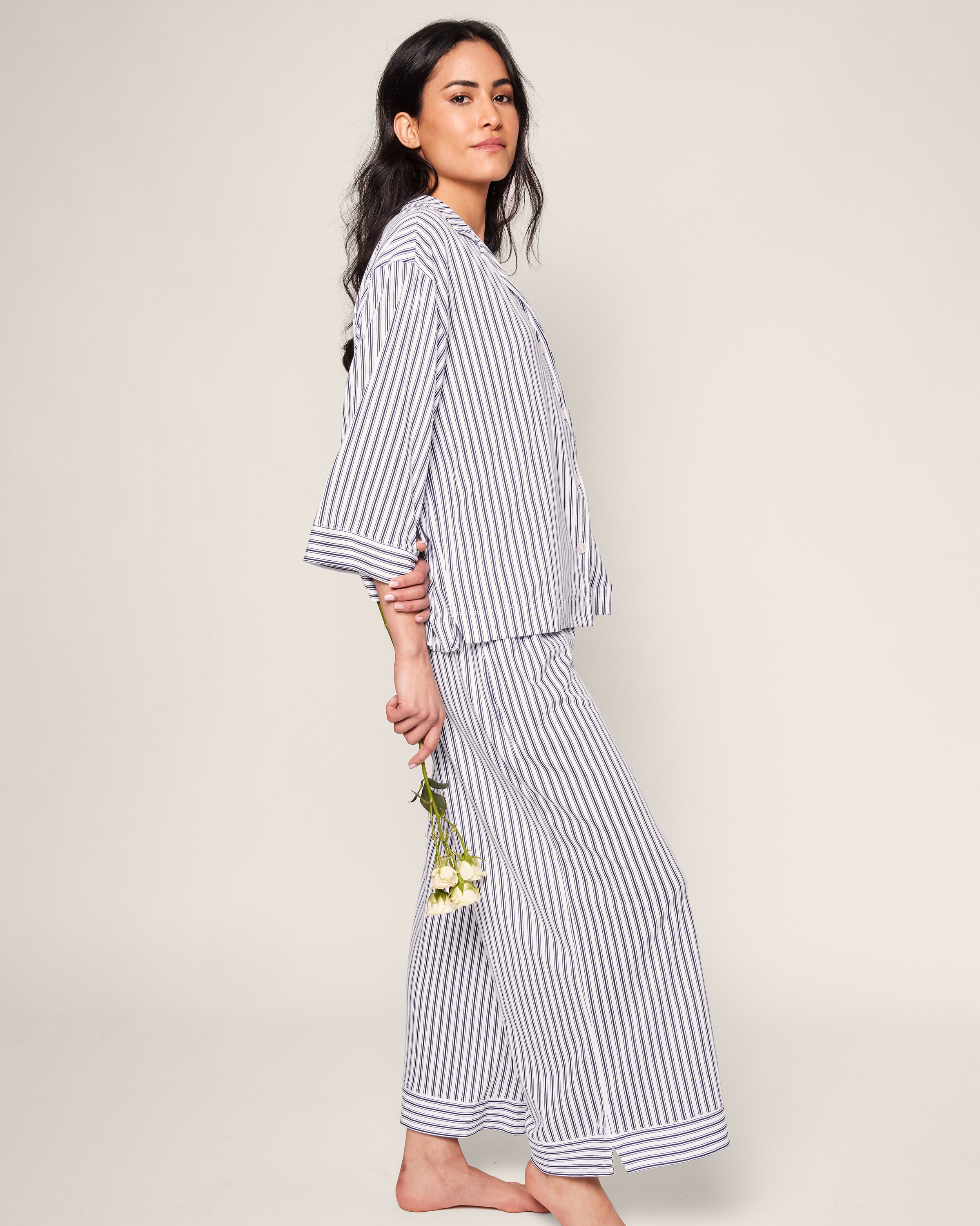 A person with long dark hair wears Petite Plumes Olivia Wide Leg Pima Pajama Set in Navy French Ticking. Barefoot, they hold a small flower bouquet behind their back against a neutral background.