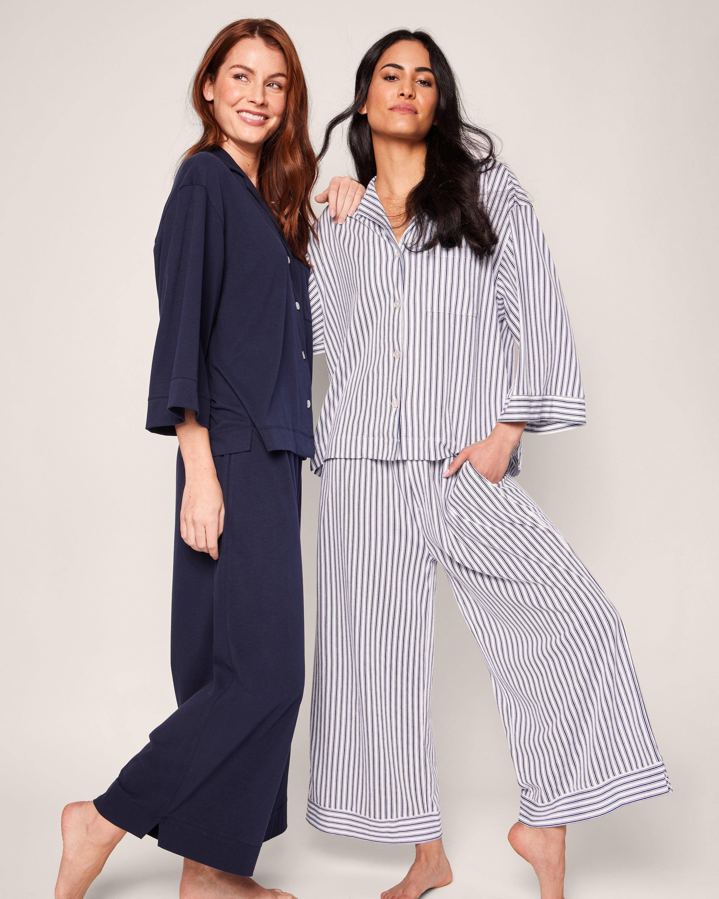 Two people pose in cozy loungewear by Petite Plume. One sports The Olivia Wide Leg Pima Pajama Set in Navy French Ticking, while the other is in a light blue and white striped Peruvian Pima cotton set. They are barefoot and smiling against a plain light background.