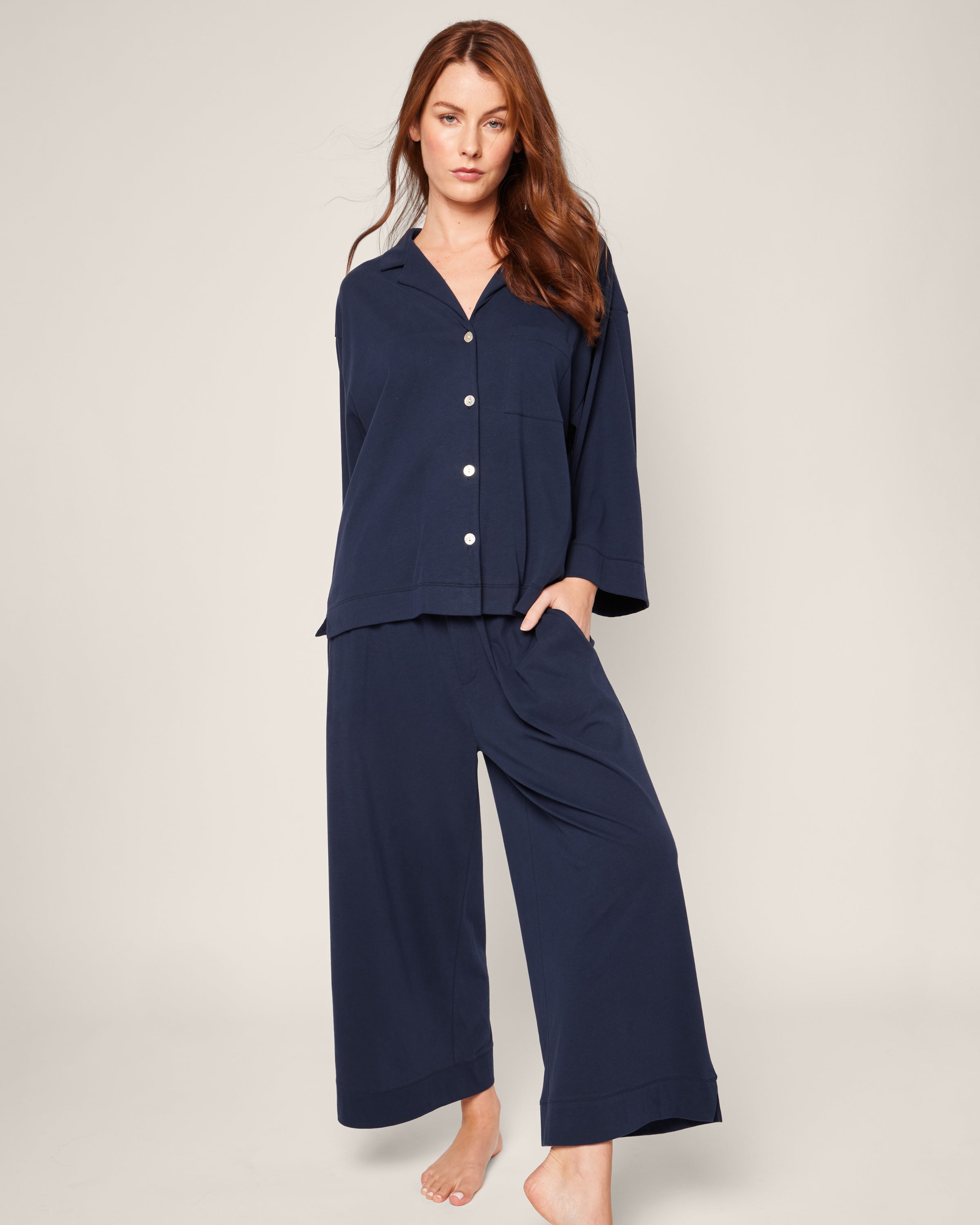 A person with long brown hair wears The Olivia Wide Leg Pima Pajama Set in Navy by Petite Plume, featuring a luxurious, buttoned, long-sleeved top and elegant wide-legged pants. They stand barefoot against an off-white background.