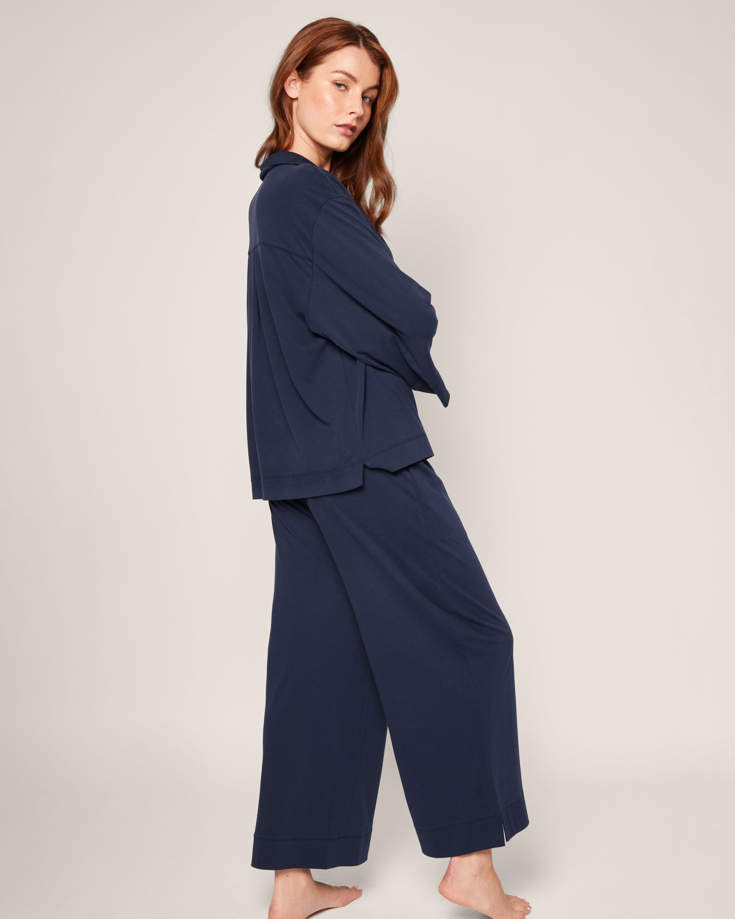 A person with long hair stands barefoot against a plain background, wearing The Olivia Wide Leg Pima Pajama Set in Navy by Petite Plume—a loose-fitting, long-sleeved top and matching wide-leg pants. They turn slightly sideways, arms crossed, radiating effortless elegance.