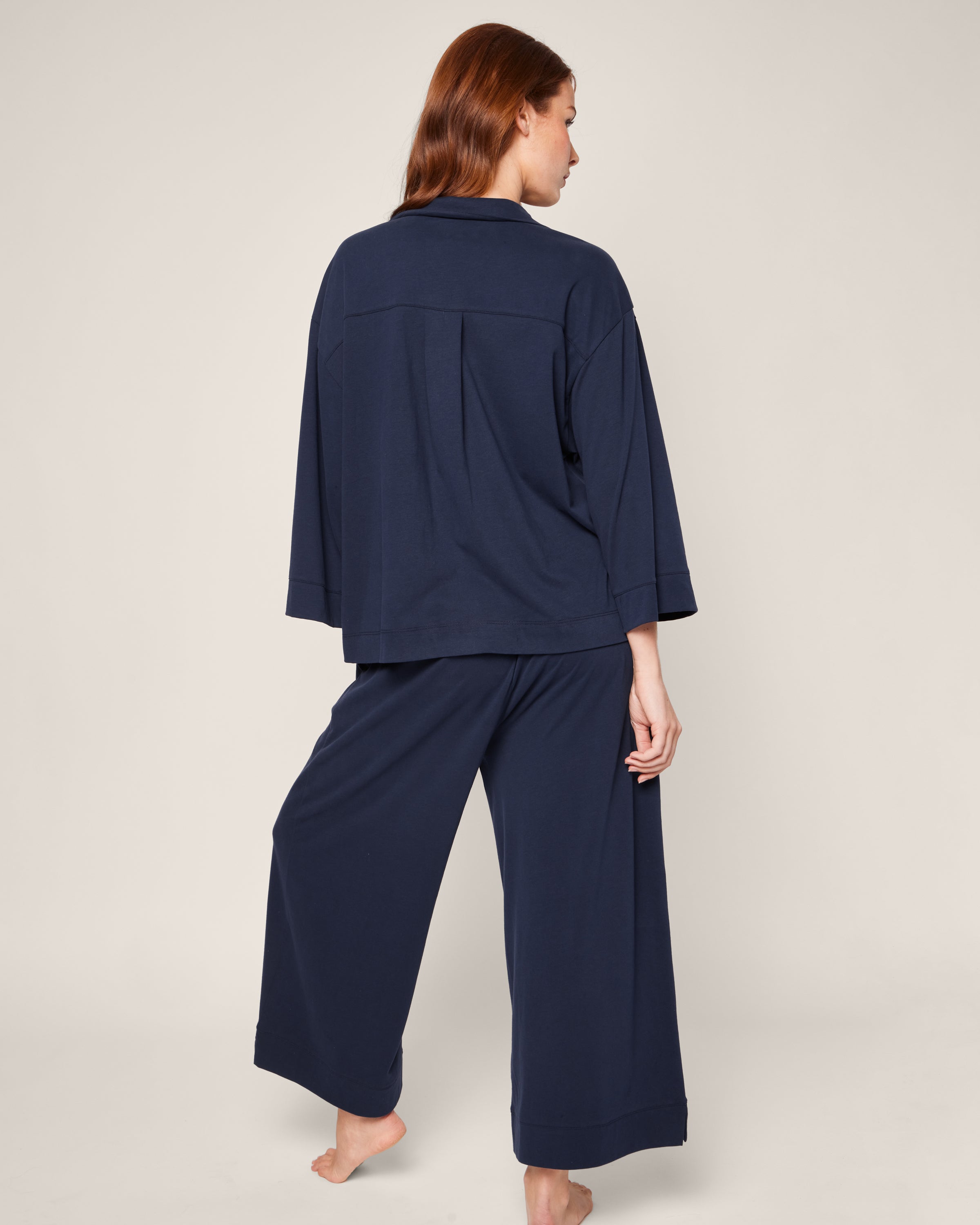 A person with long brown hair stands barefoot, facing away from the camera in The Olivia Wide Leg Pima Pajama Set in Navy by Petite Plume. The luxurious, loose-fitting pants and long-sleeved top offer an elegant sleepwear look against a plain off-white background.