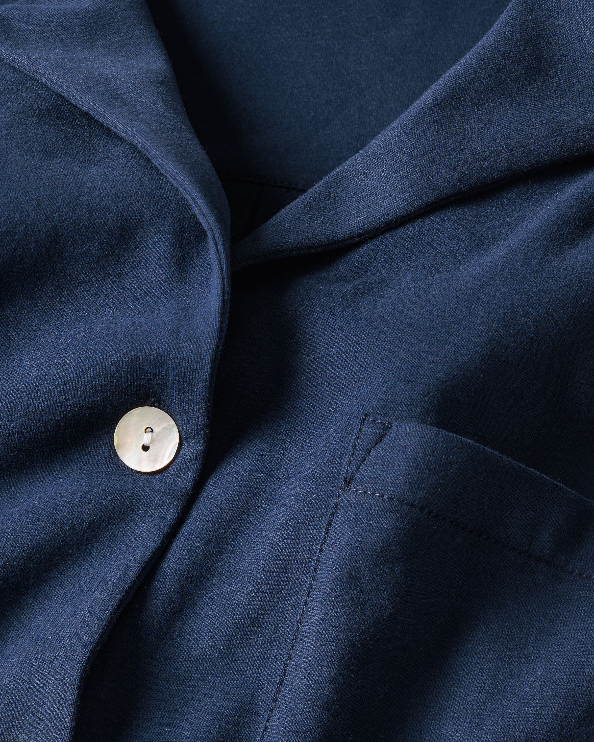 A close-up of The Olivia Wide Leg Pima Pajama Set in Navy by Petite Plume showcases its soft, smooth fabric, a lapel, and single metallic button with two holes. A visible right pocket enhances the elegantly tailored design reminiscent of luxury sleepwear.