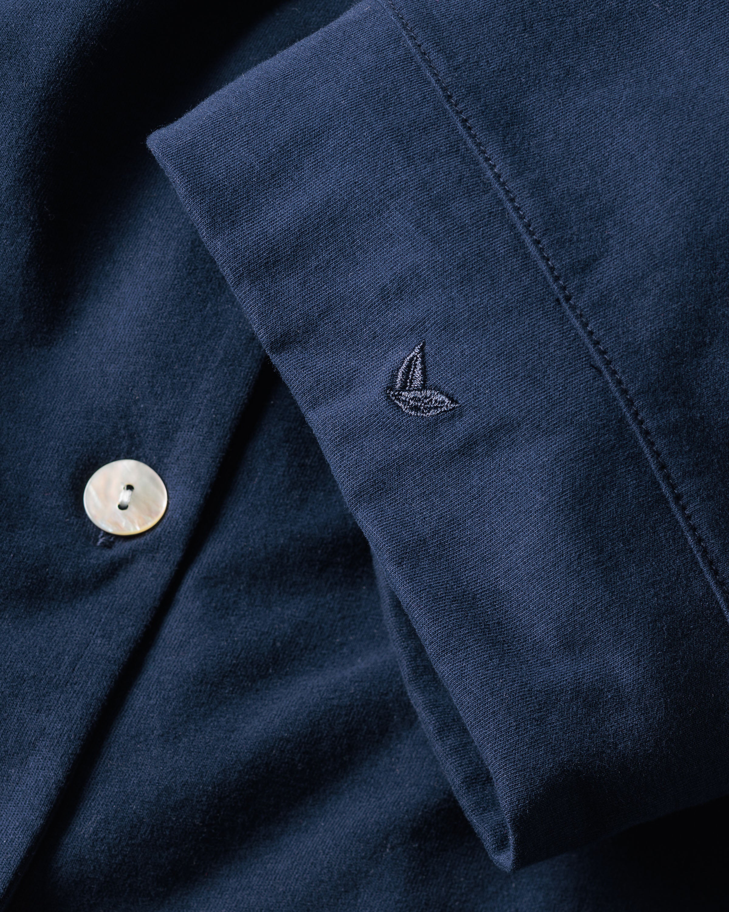 Close-up of The Olivia Wide Leg Pima Pajama Set in Navy by Petite Plume, with folded dark blue fabric featuring a small embroidered leaf logo and a pearl button on the left, exemplifying exquisite luxury sleepwear craftsmanship.