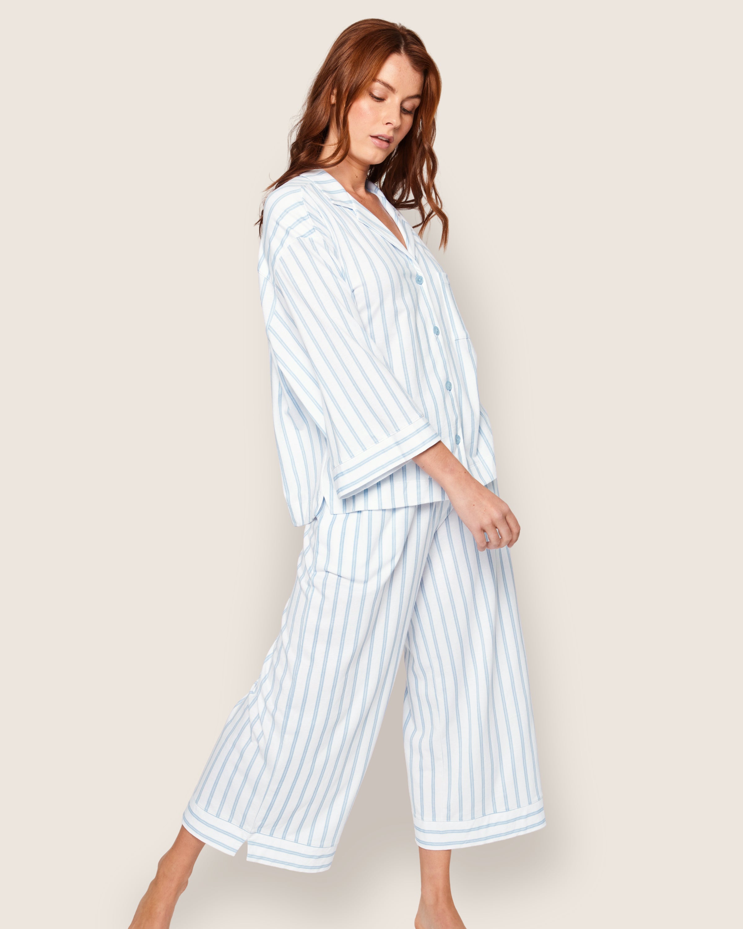 A person poses gracefully in the Olivia Wide Leg Pima Pajama Set by Petite Plume. This luxurious, loose-fitting ensemble, featuring a button-up shirt and wide-legged pants in periwinkle stripe, stands out against a neutral background.