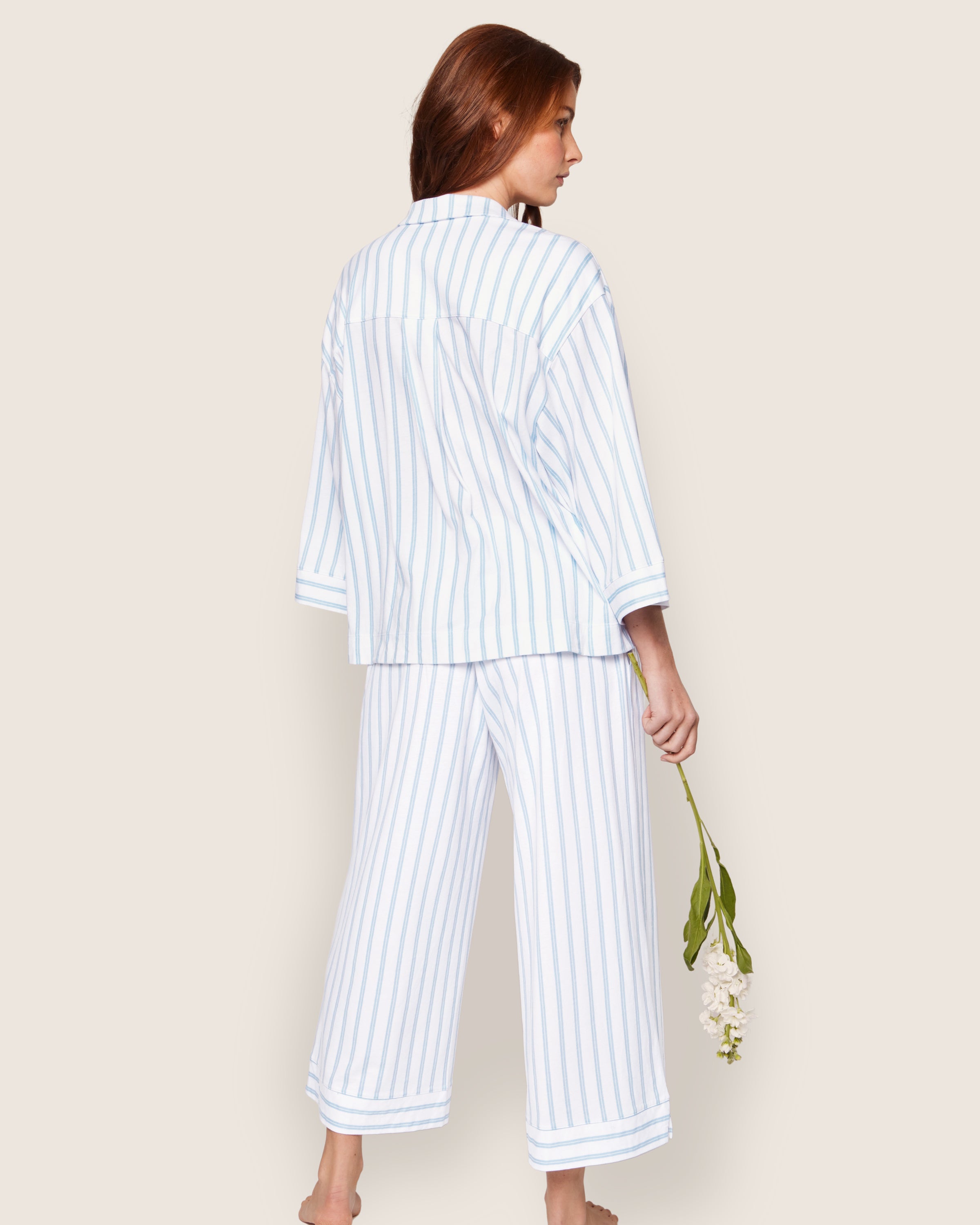 Standing barefoot on a light background, a person with long hair is wrapped in Petite Plumes luxurious Olivia Wide Leg Pima Pajama Set in Periwinkle Stripe, holding a white flower and glancing over their shoulder with effortless elegance.