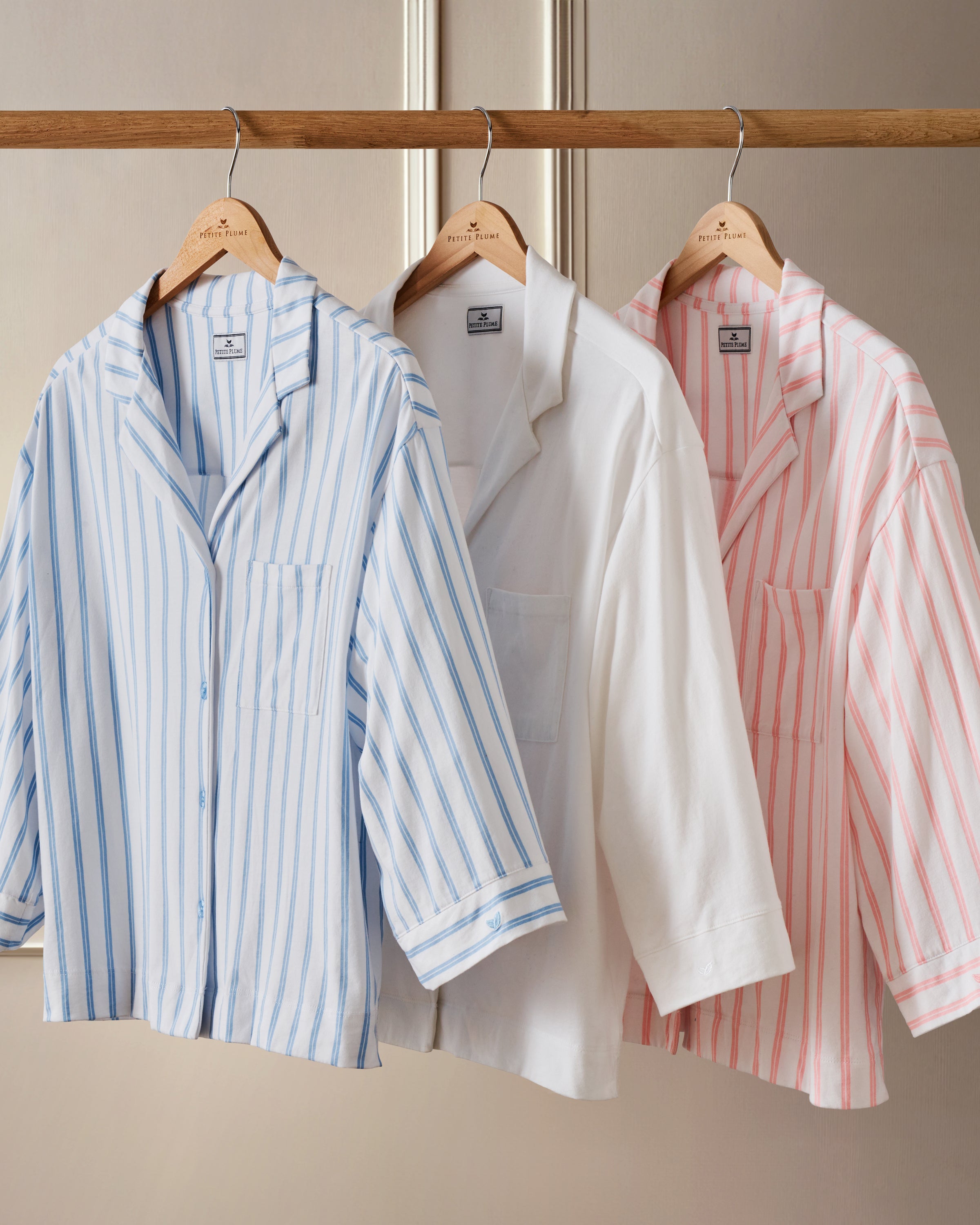 Three shirts with collars, front pockets, and buttons are on wooden hangers against a beige background. Made from breathable fabric, they include a blue striped style like The Olivia Wide Leg Pima Pajama Set in Periwinkle Stripe by Petite Plume, exuding elegant sleepwear charm for everyday use.