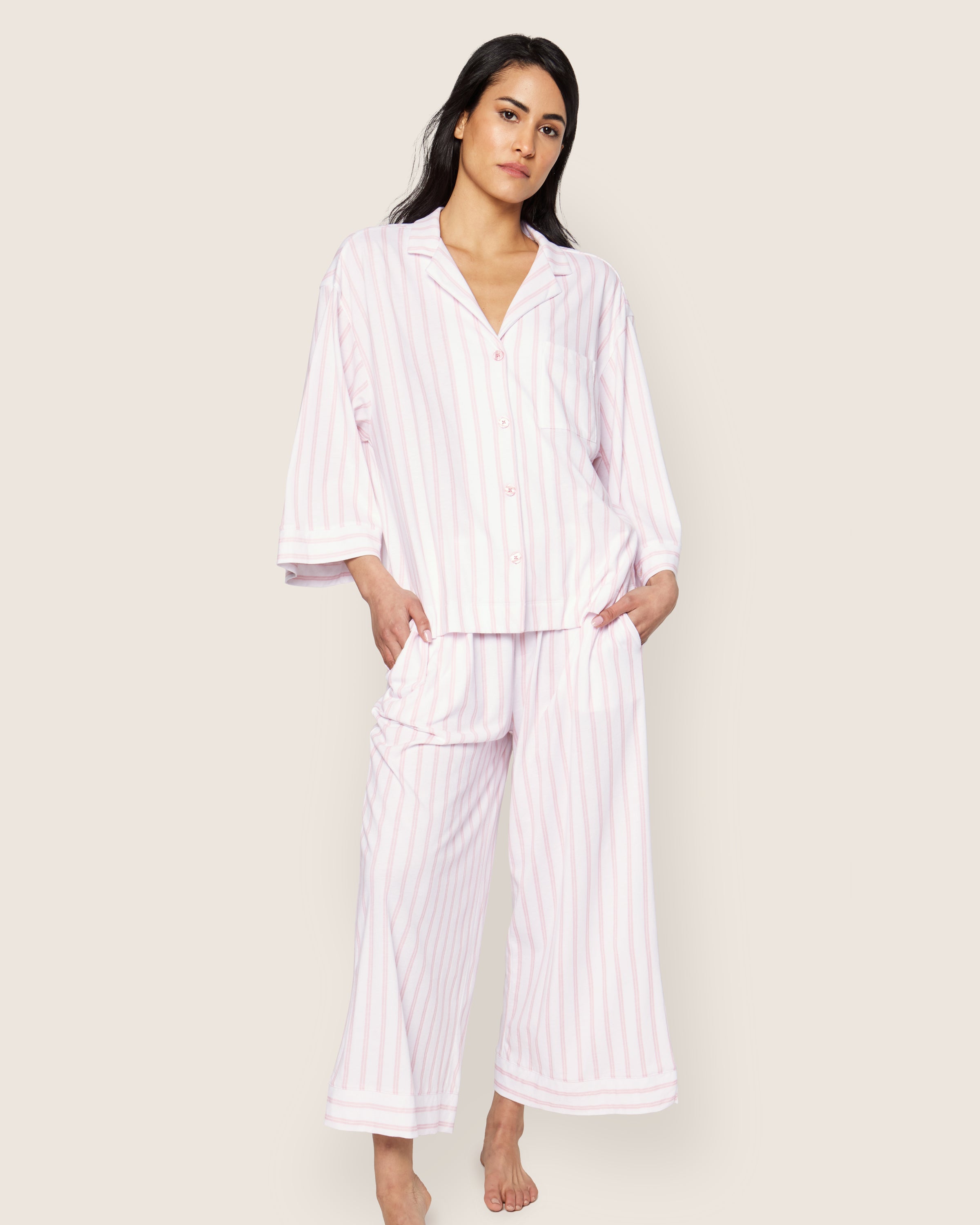 A person is seen in The Olivia Wide Leg Pima Pajama Set in Pink Stripe by Petite Plume, featuring a button-up top with a collar and long sleeves, and wide-leg pants made of luxurious Peruvian Pima cotton. The backdrop is plain and light-colored.