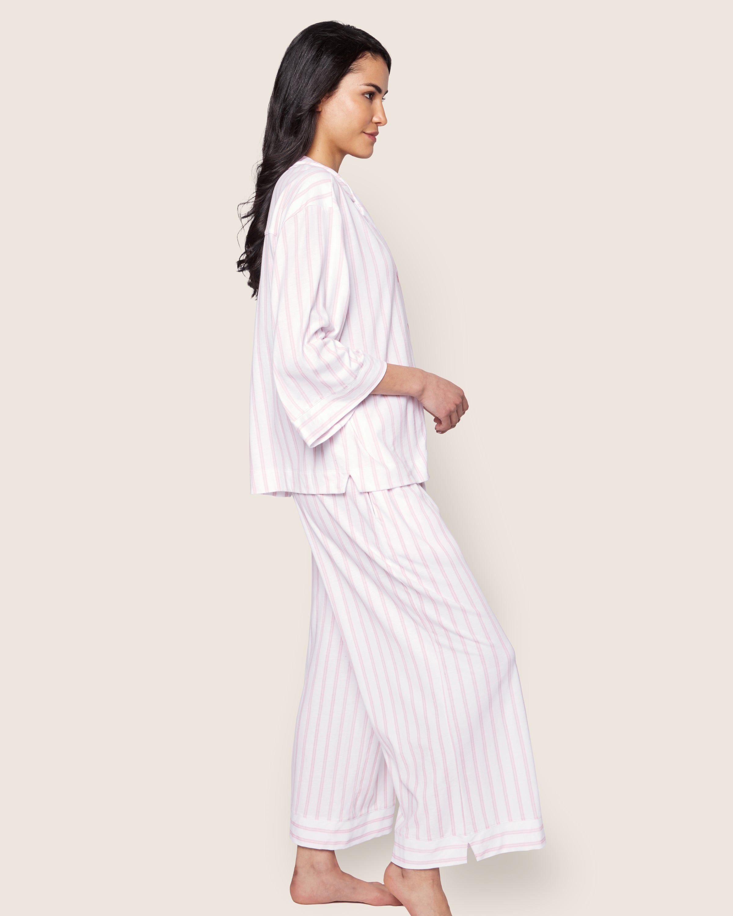 A person with long dark hair stands barefoot in The Olivia Wide Leg Pima Pajama Set in Pink Stripe by Petite Plume, featuring a soft Peruvian Pima cotton long-sleeve top and wide-leg pants. The background is a plain light color.