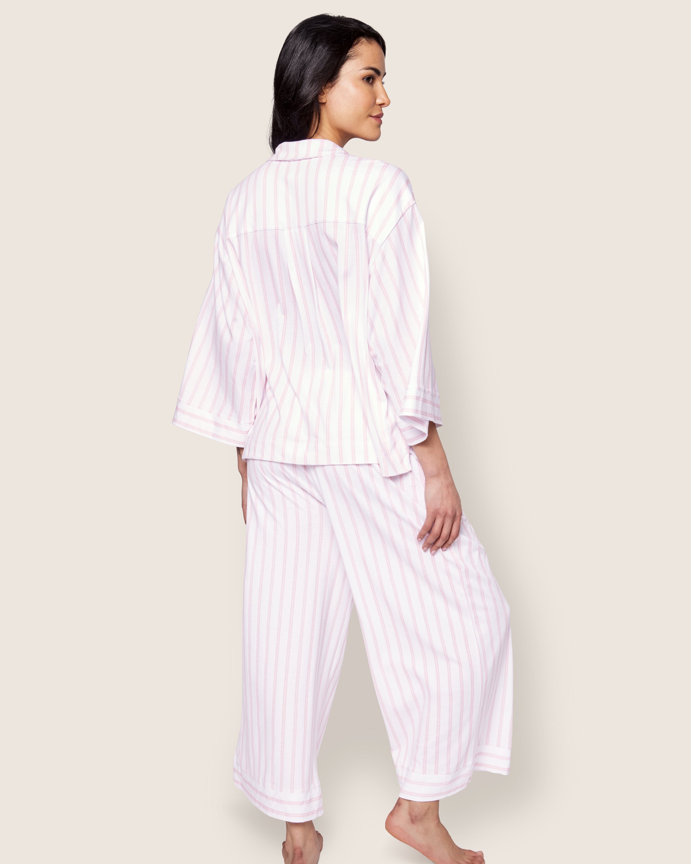 Someone barefoot, standing on one leg and looking to the side, captures a serene bedtime ritual against a light background. They are wearing Petite Plumes The Olivia Wide Leg Pima Pajama Set in Pink Stripe, crafted from luxurious Peruvian Pima cotton with a long-sleeve top and wide-leg pants.