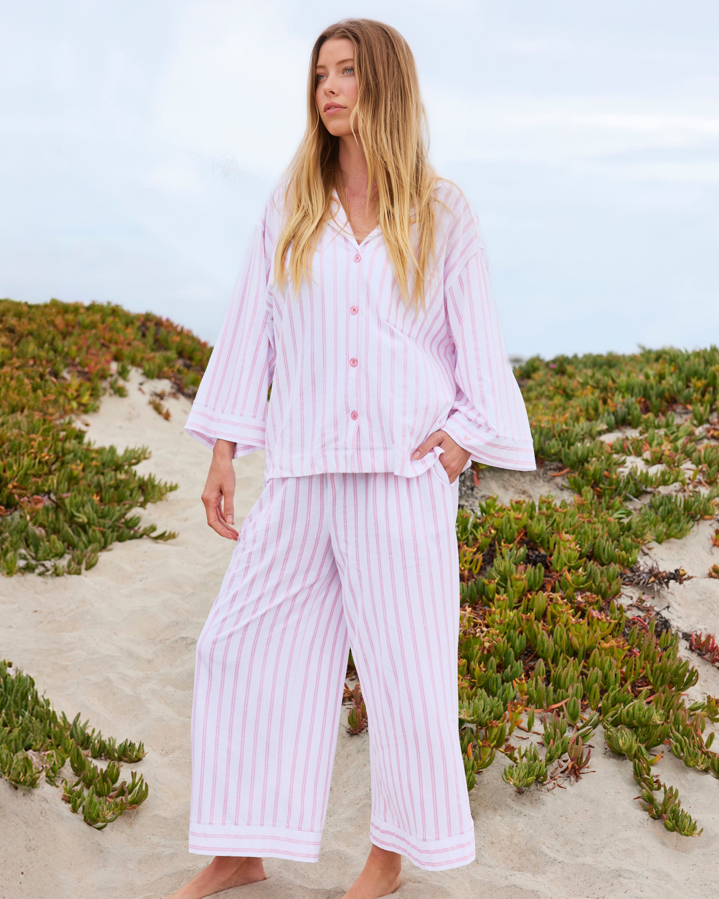 On a sandy beach with lush greenery, a person stands barefoot in The Olivia Wide Leg Pima Pajama Set in Pink Stripe from Petite Plume, made of soft Peruvian Pima cotton, under an overcast sky.