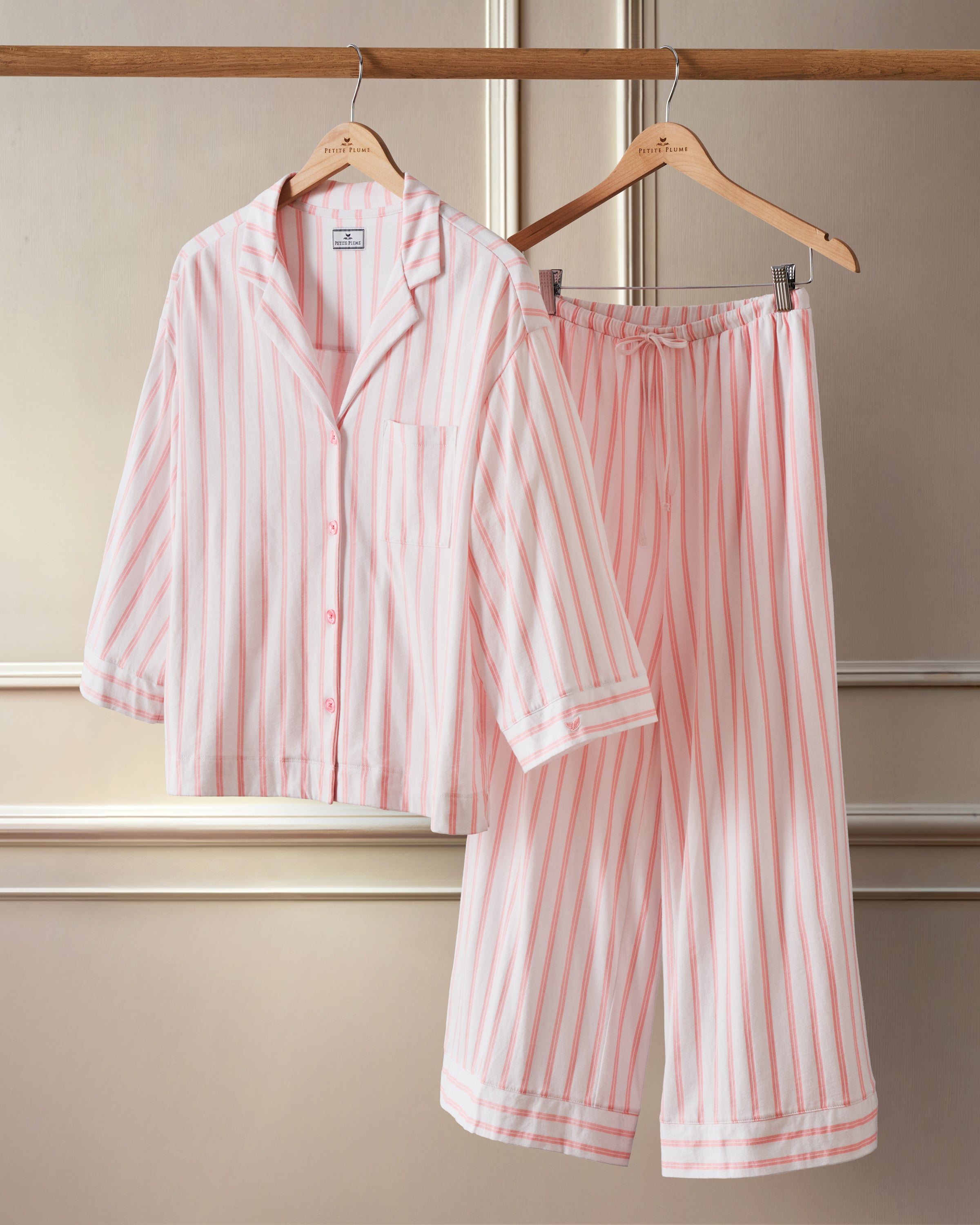 The Olivia Wide Leg Pima Pajama Set in Pink Stripe by Petite Plume, crafted from soft Peruvian Pima cotton, featuring a button-up shirt and elastic-waist pants, hangs on wooden hangers against a cream paneled wall.