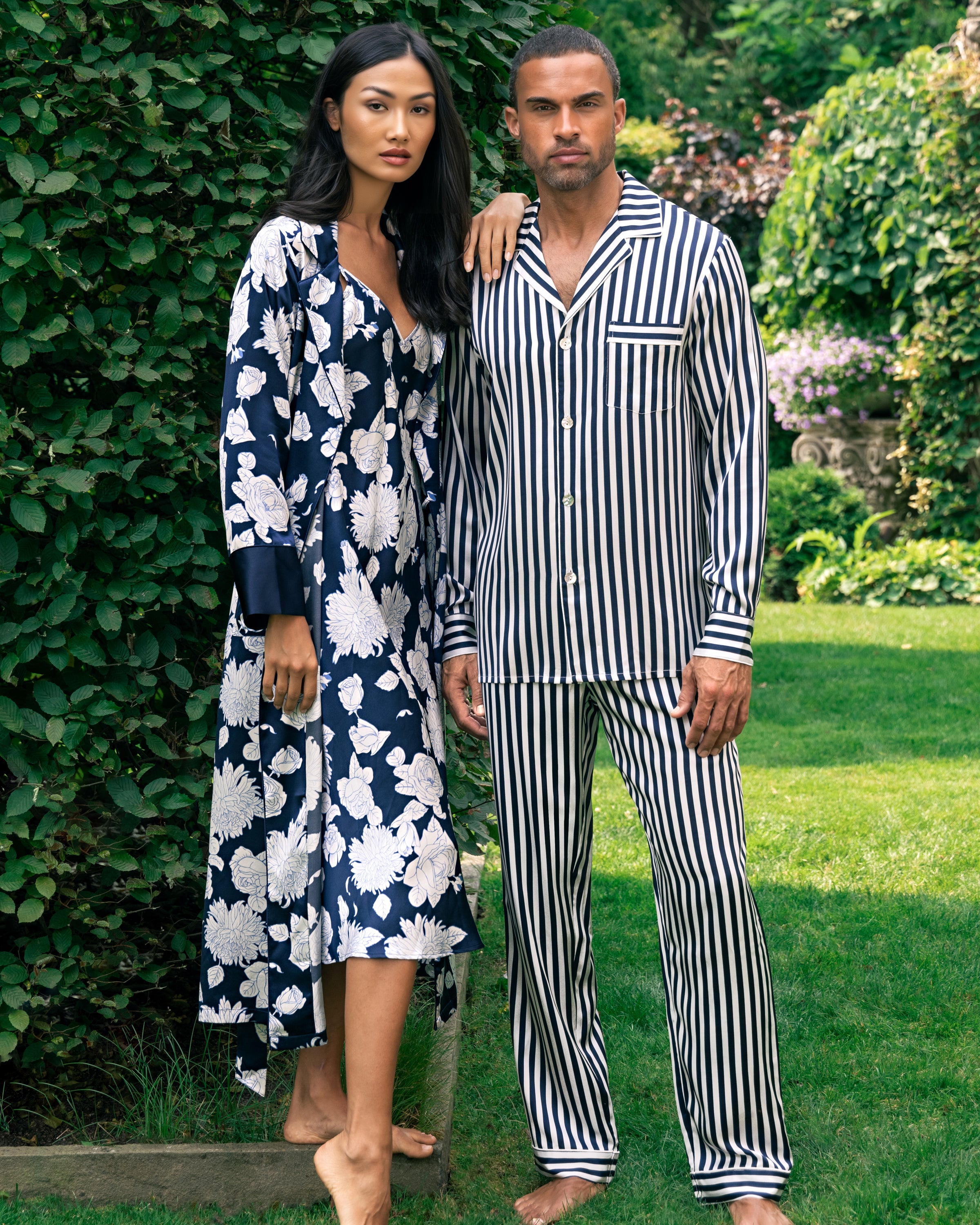 A woman stands barefoot on grass in a long, floral-patterned robe beside a man in the Mens Silk Pajama Set in Navy Bengal Stripe by Petite Plume. They are next to a tall hedge with a lush garden behind them.