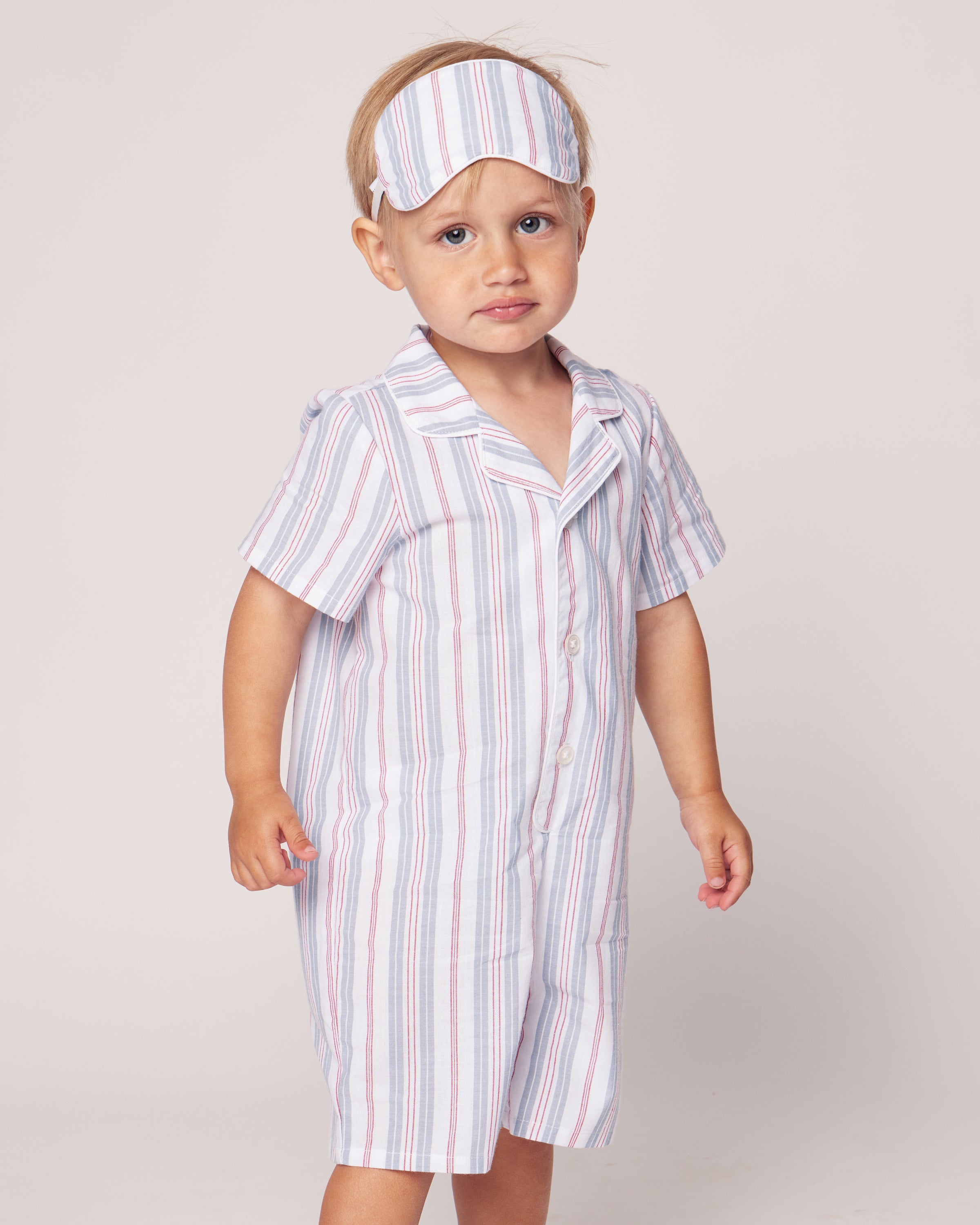 A young child in Petite Plumes Babys Twill Summer Romper with Vintage French Stripes, paired with a sleep mask, stands against a plain background looking slightly puzzled. The luxurious romper features pale blue, pink, and white stripes.