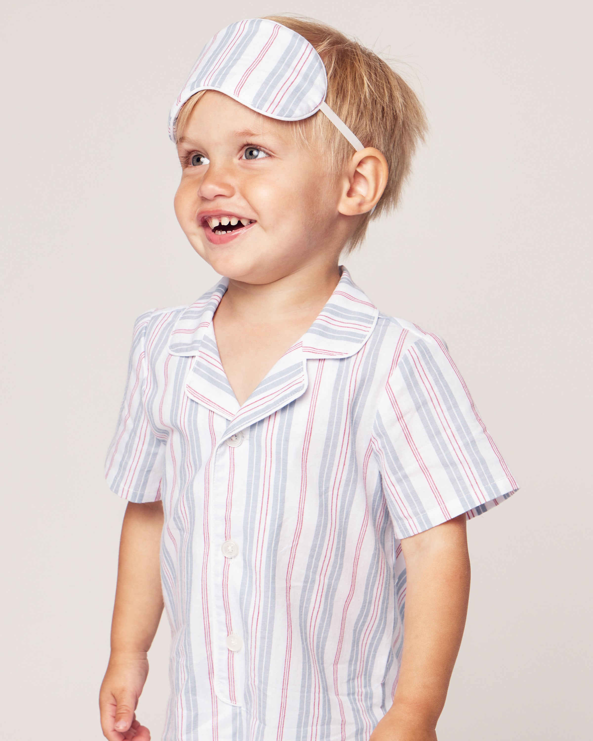 A smiling young child in Petite Plumes luxurious Babys Twill Summer Romper in Vintage French Stripes, with matching light blue and pink hues and a sleep mask on their head, gazes to the side against a plain background.