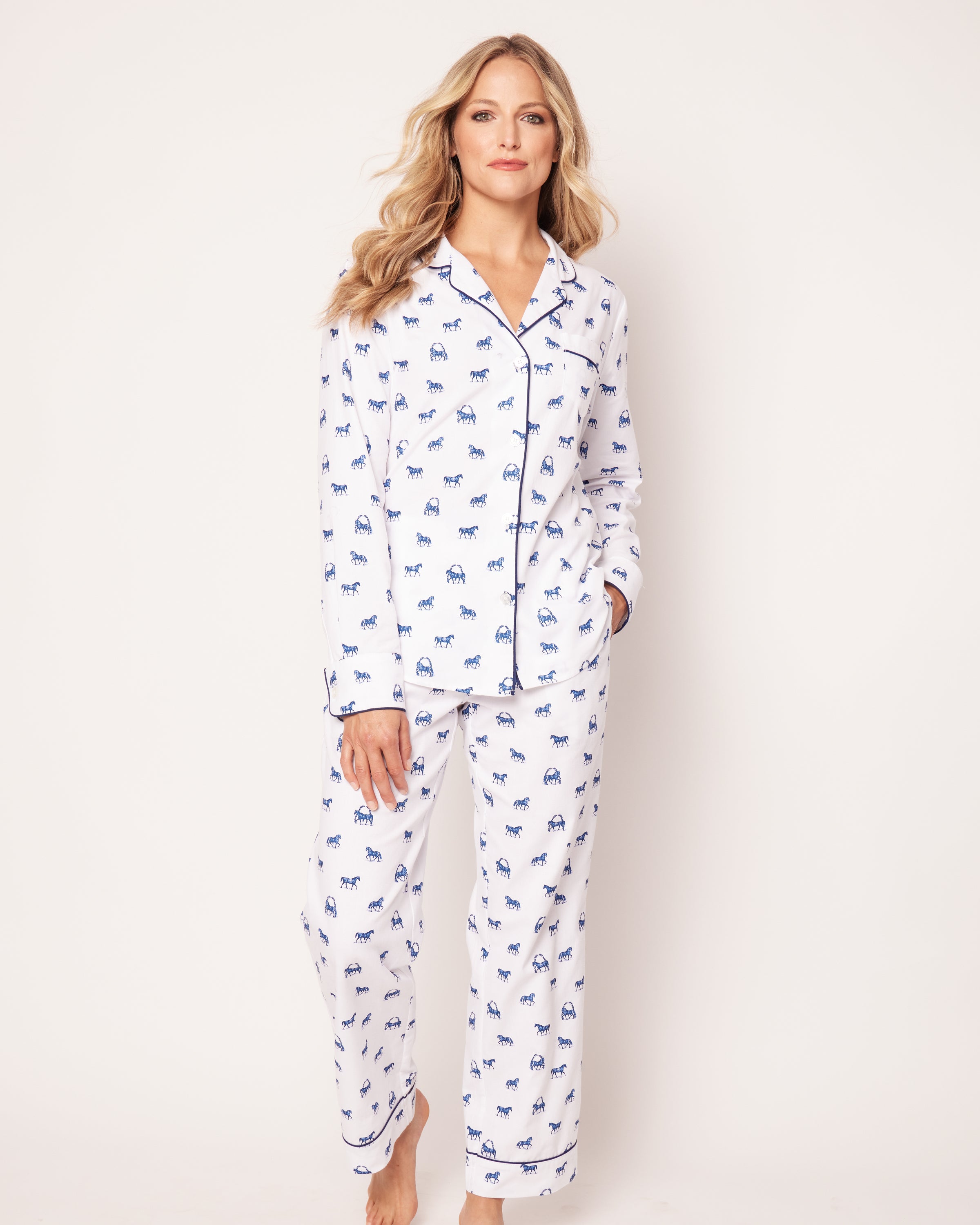 A person with long, wavy hair poses against a plain backdrop wearing Petite Plumes Womens Twill Pajama Set in The Equestrian. The set features a fine cotton collared, long-sleeve top with dark piping and matching pants adorned with an elegant blue elephant print.