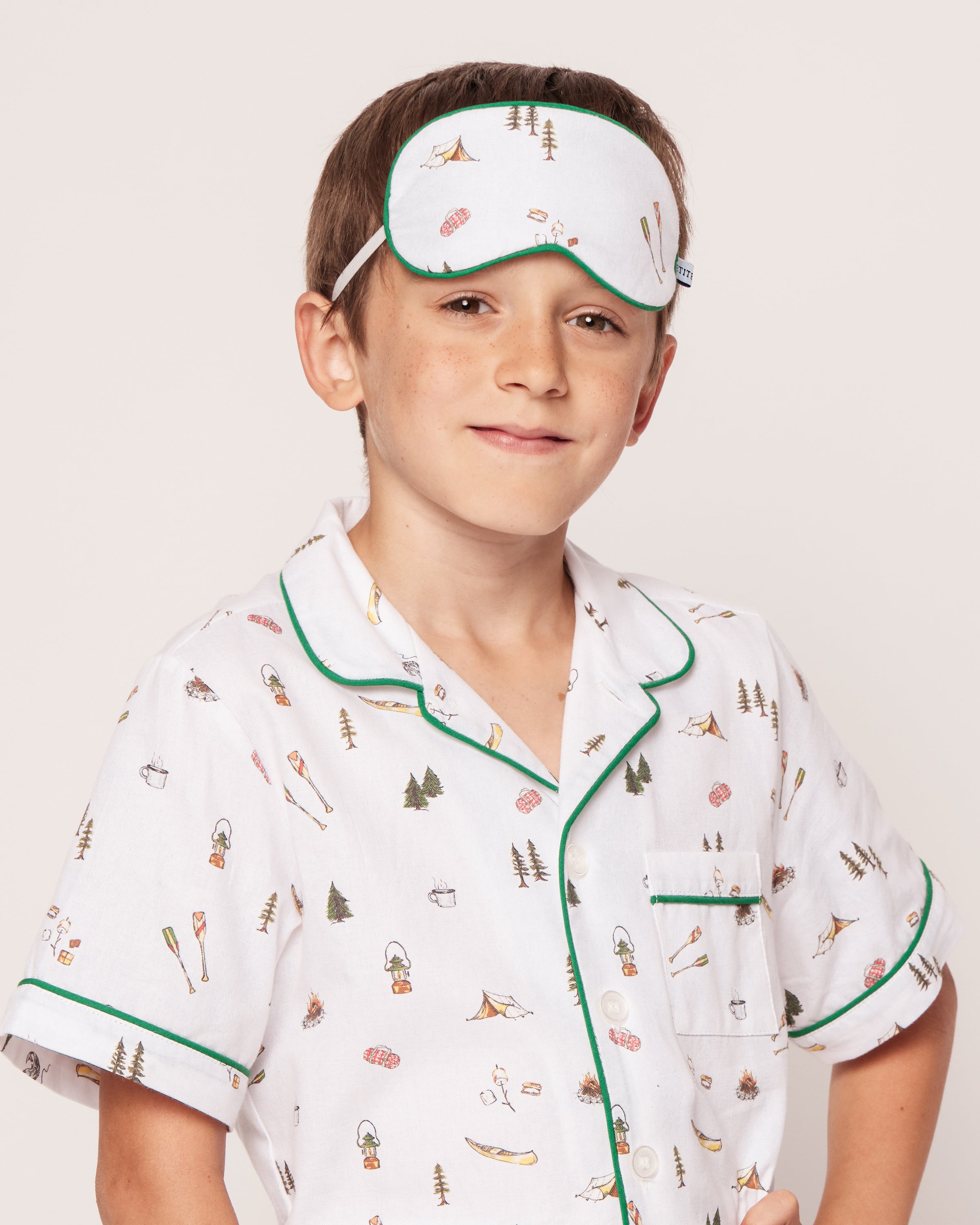 A young boy in Petite Plumes Kids Sleep Mask in The Great Outdoors stands confidently, wearing white pajamas with small illustrations and green piping. With a matching eye mask on his forehead and a slight smile, he poses with his hand on his hip against a light-colored plain background.