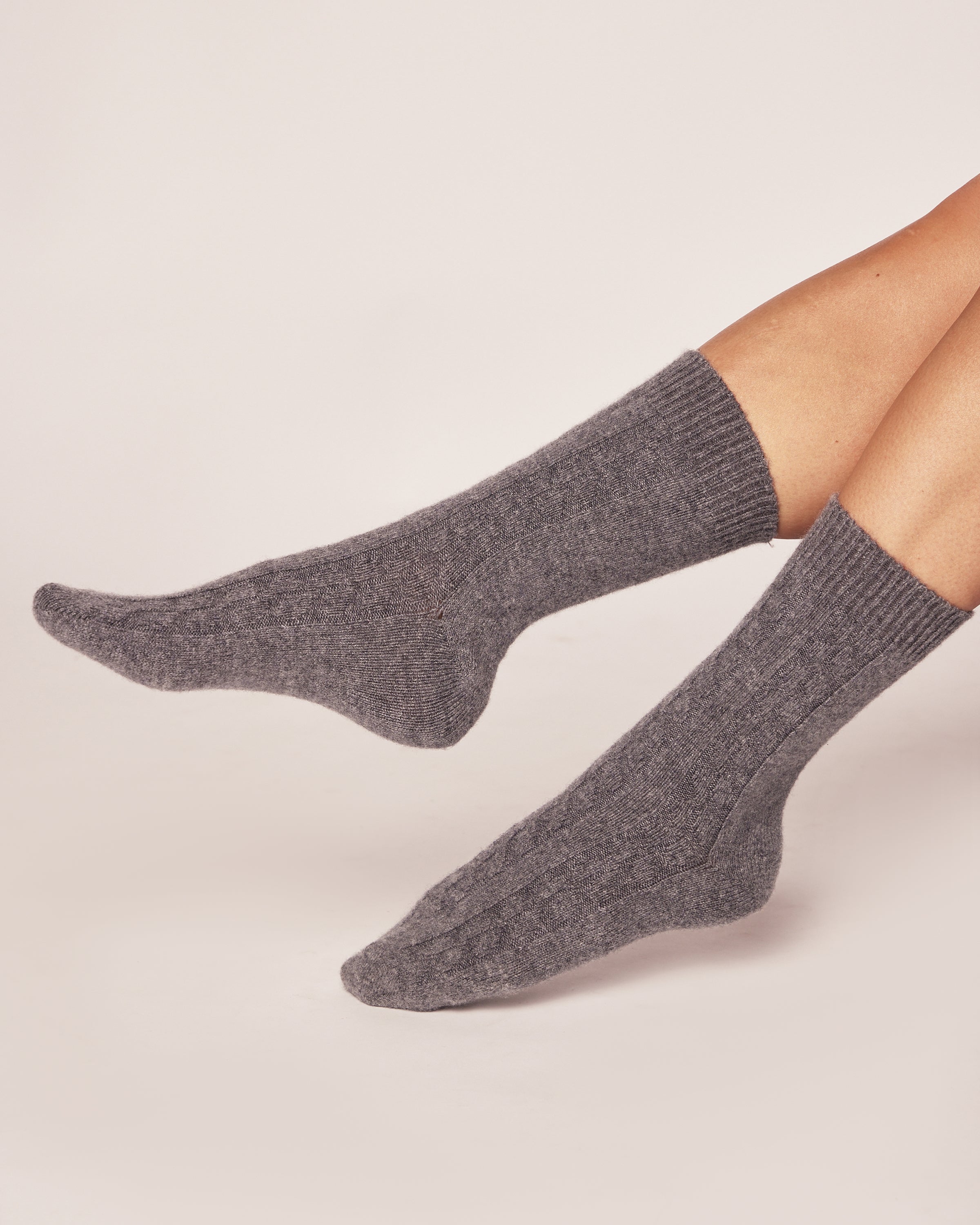 Two feet are adorned with cozy, textured Petite Plume Womens Cashmere Socks in Dark Grey. The plain background highlights their snug fit and softness. Perfect for gifting, they come elegantly packaged in a stylish gift box.