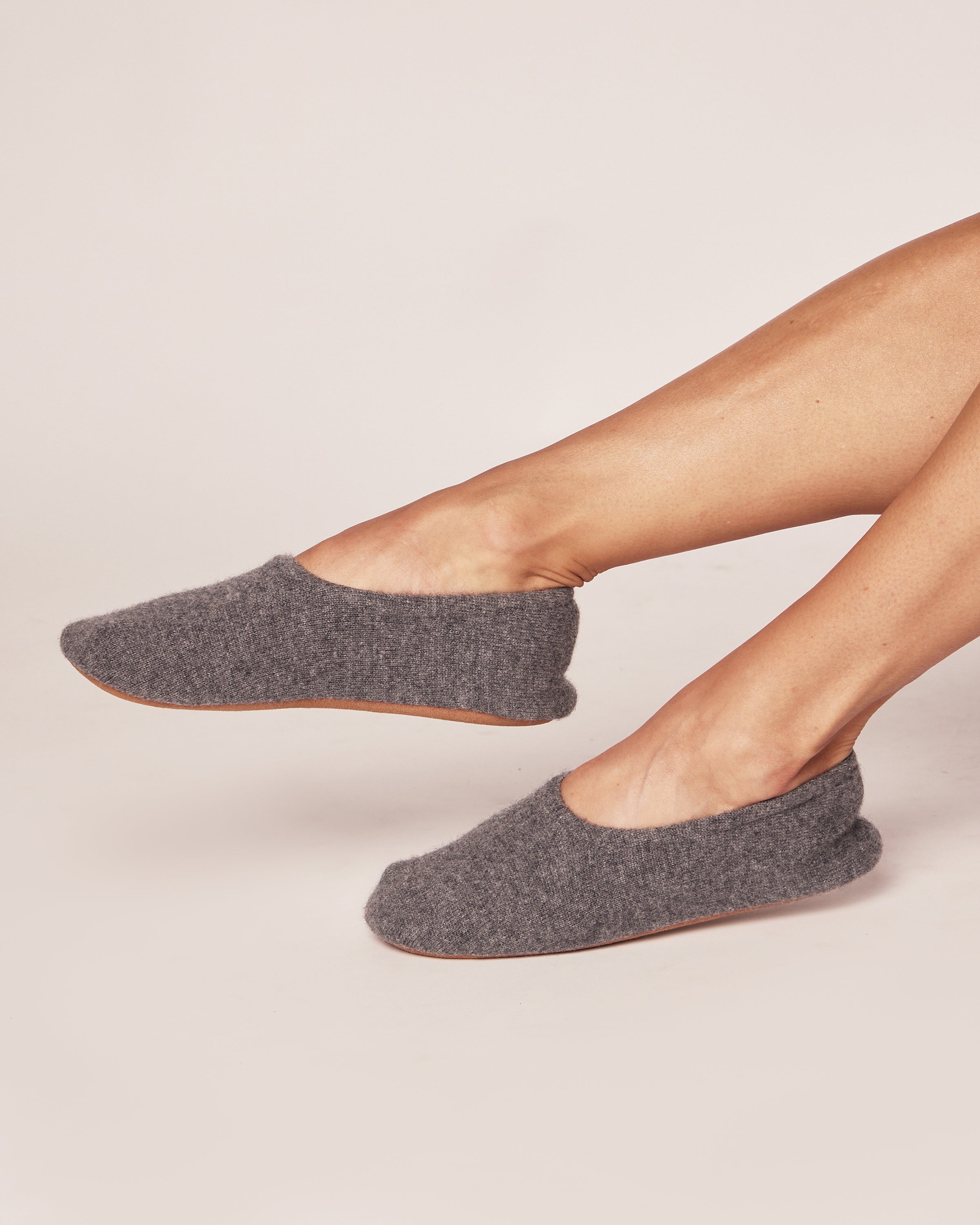 A person wearing Petite Plumes Womens Cashmere Slippers in Dark Grey with tan soles stretches their legs against a white backdrop, highlighting the comfort of the luxurious cashmere blend.