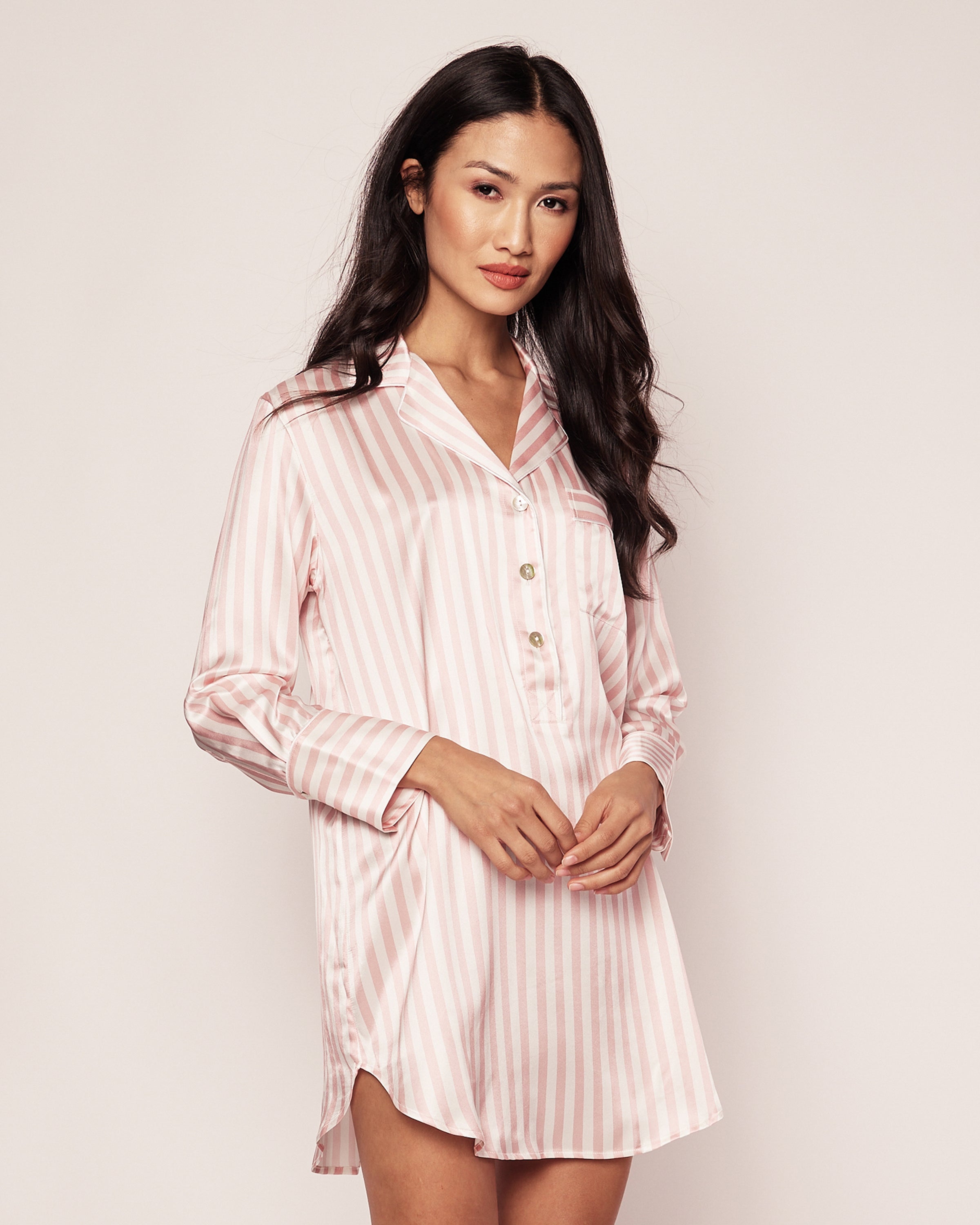 A person poses against a plain background in the Womens Silk Nightshirt in Pink Stripe by Petite Plume. The pink and white striped button-up shirt, reminiscent of cozy sleepwear, has a loose fit and collar. The persons hands are gently clasped in front.