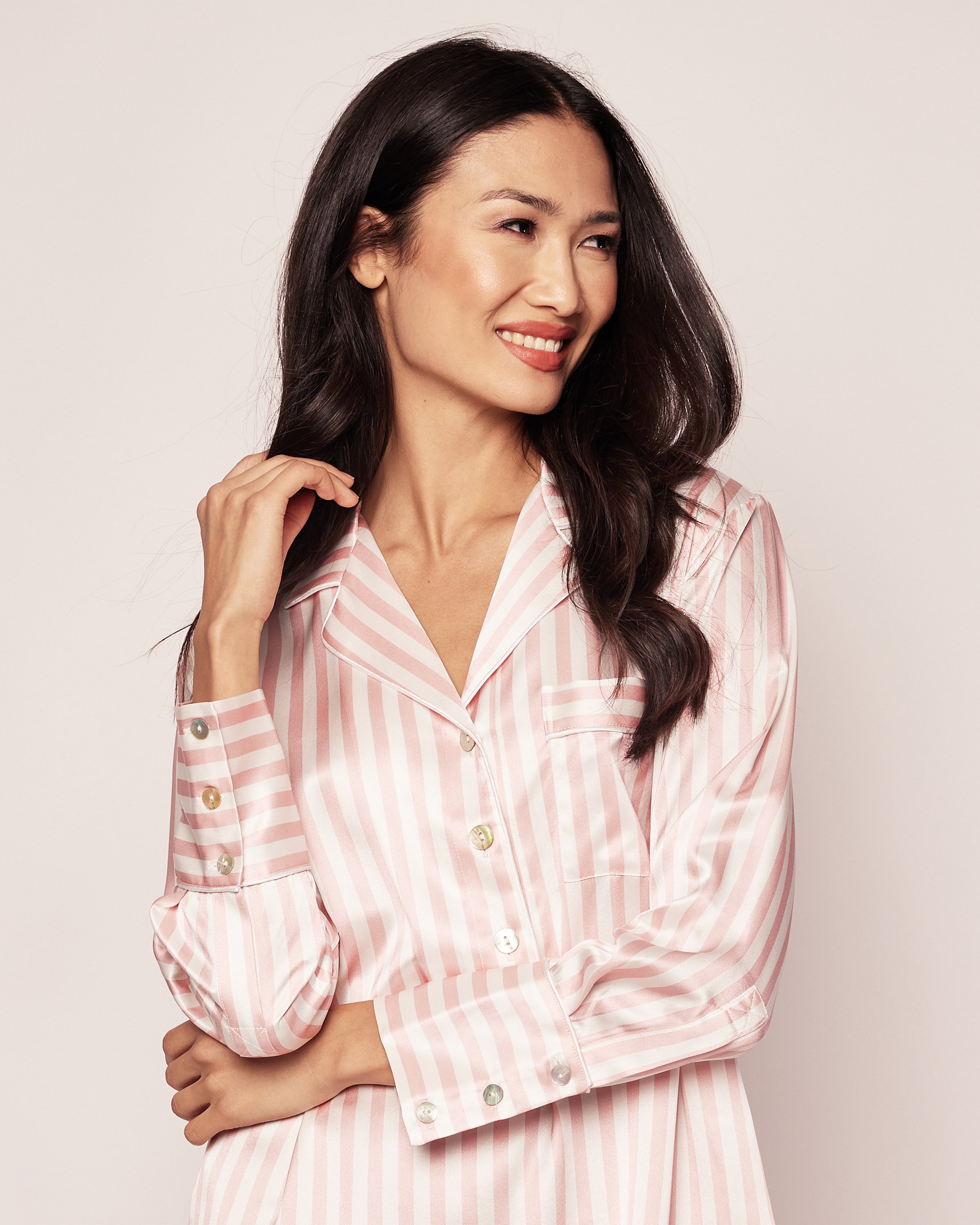 A woman with long dark hair smiles while wearing Petite Plumes Womens Silk Nightshirt in Pink Stripe. She gently touches her hair with one hand against a plain, light-colored background.