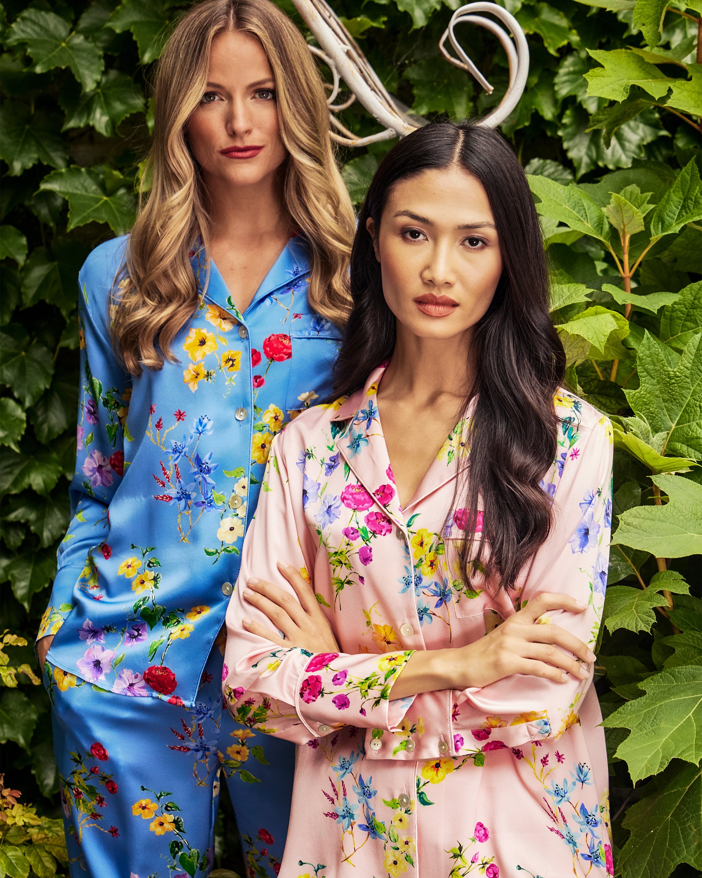 Two women with long hair stand in a lush garden wearing luxurious floral pajamas. The woman on the left wears Petite Plumes Womens Silk Pajama Set in Brilliant Botanical Azure, while the other is dressed in pink botanical print pajamas, exuding elegance and comfort.