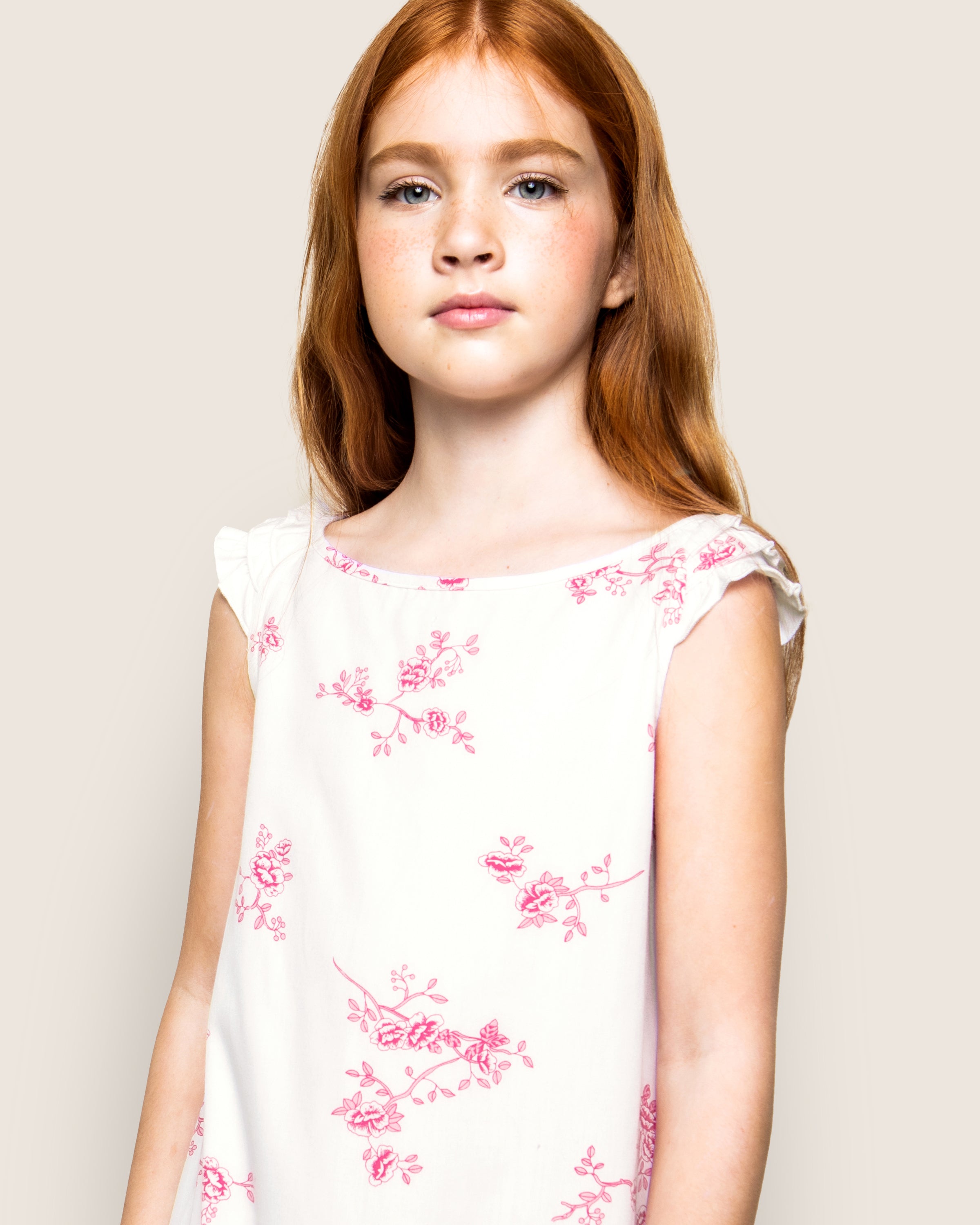 A young girl with long red hair and freckles, wearing Petite Plumes Girls Twill Amelie Nightgown in English Rose Floral, stands against an off-white background with a calm expression.