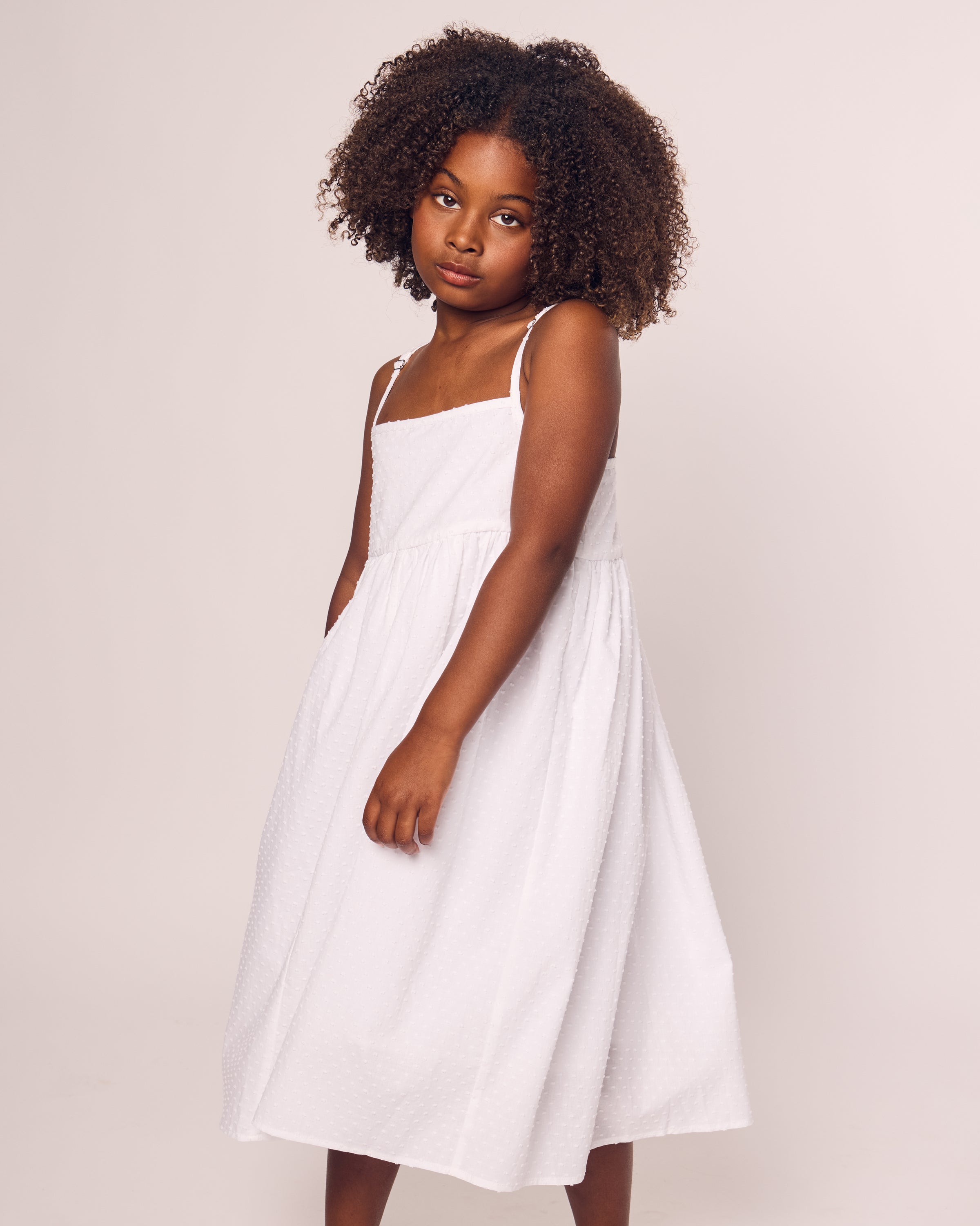 A young girl with curly hair stands against a neutral background, wearing the Petite Plume Girls Swiss Dots Serene Day Dress in White, made of Cotton Swiss Dot. She gazes calmly at the camera.