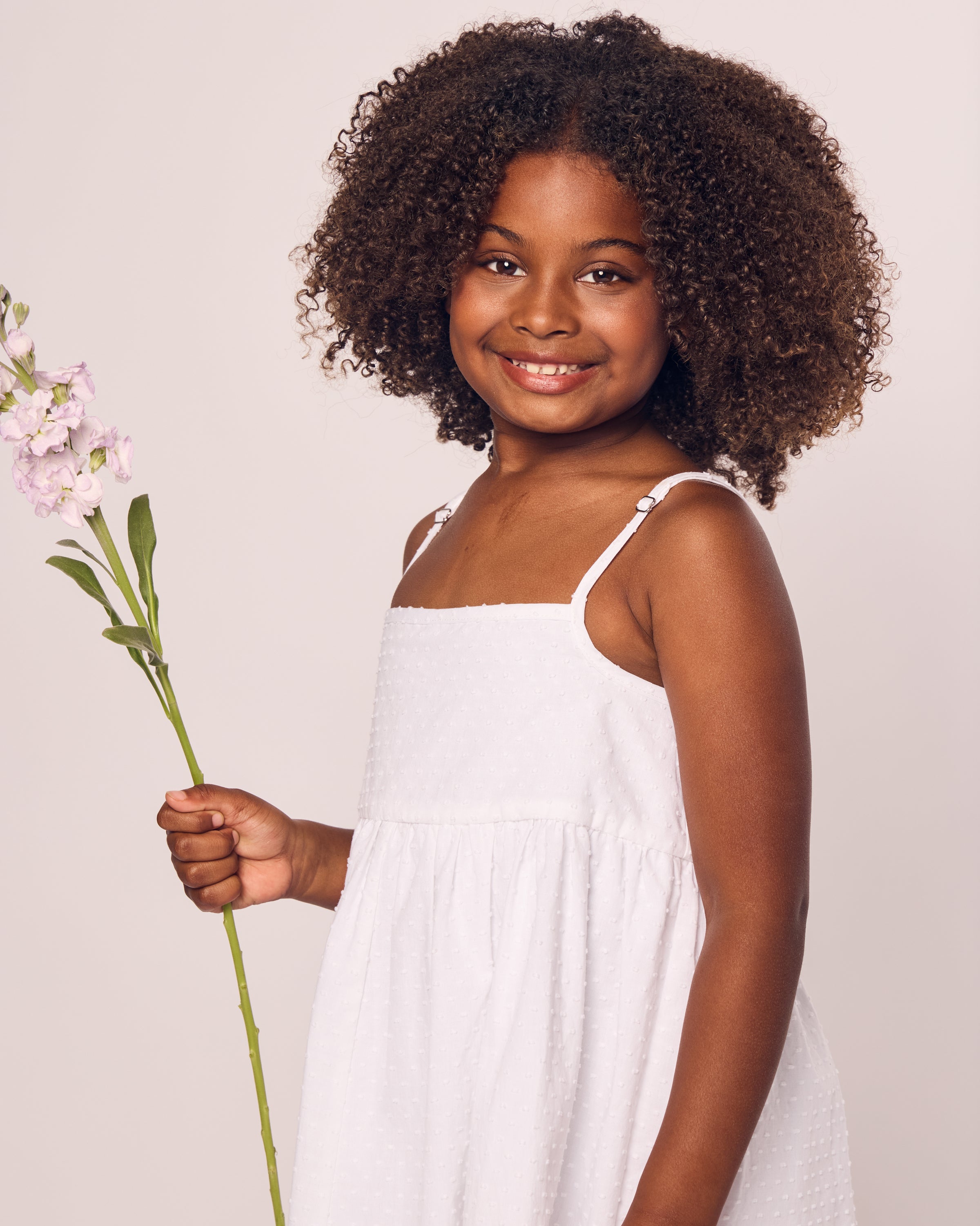 A young girl with curly hair in Petite Plumes Girls Swiss Dots Serene Day Dress in White smiles, holding a light pink flower stem. She poses gracefully against a plain backdrop, exuding innocence and charm.