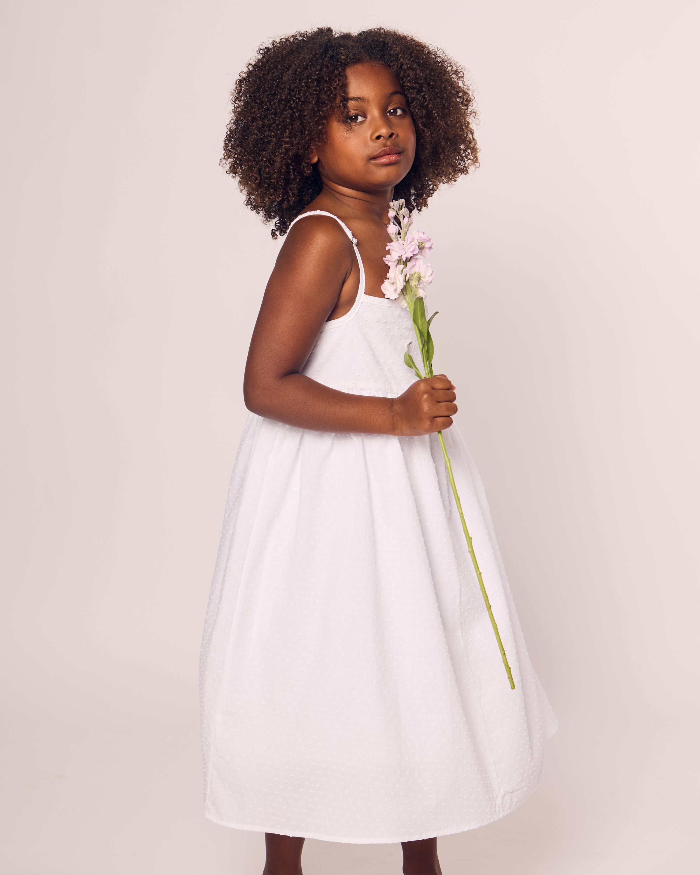 A young girl with curly hair stands in the Petite Plume Girls Swiss Dots Serene Day Dress in White, made from luxurious cotton fabric, holding a light pink flower. She gazes softly to the side against a plain background.