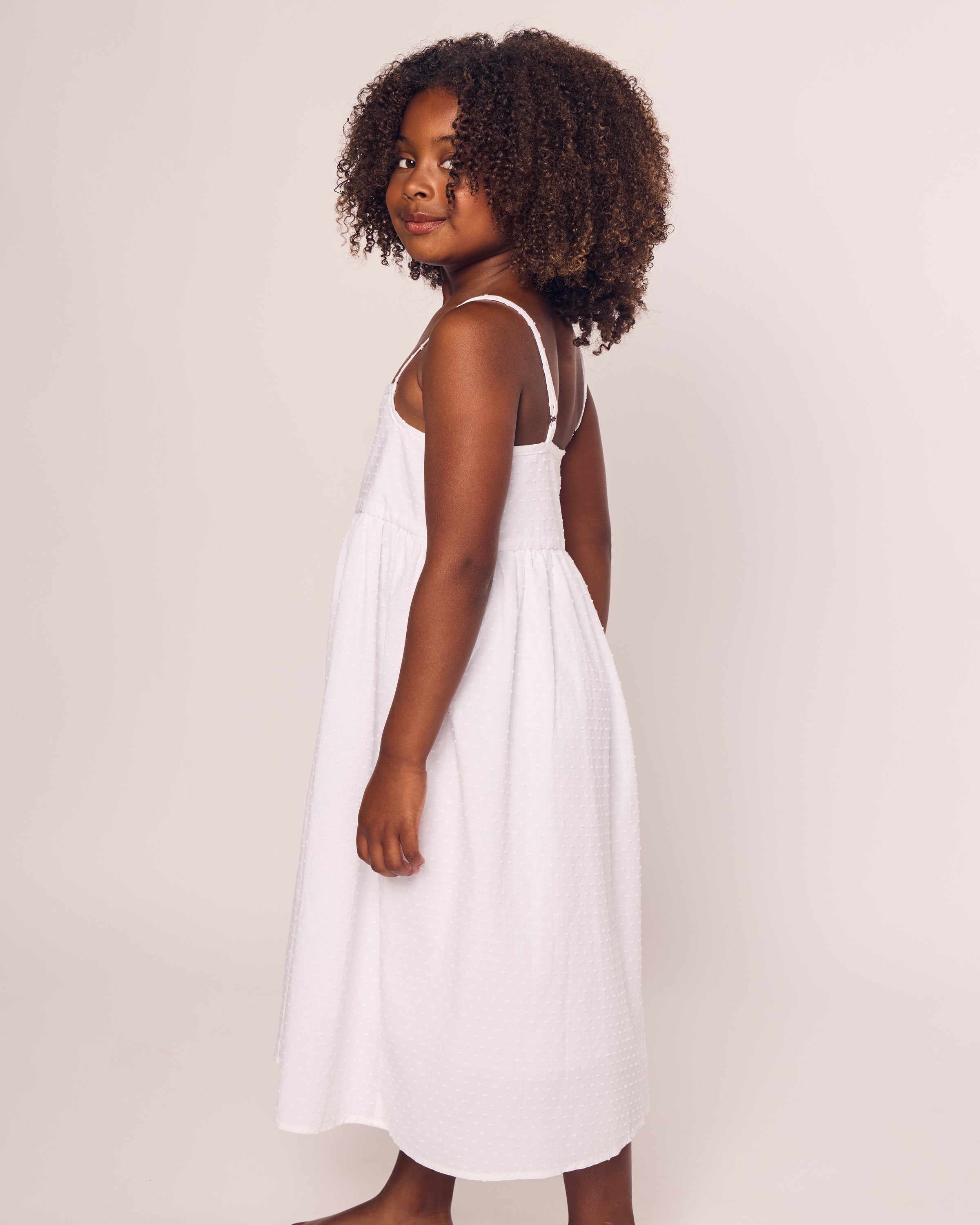 A young girl with curly hair, wearing the Petite Plume Girls Swiss Dots Serene Day Dress in White, stands sideways against a plain background, looking at the camera.