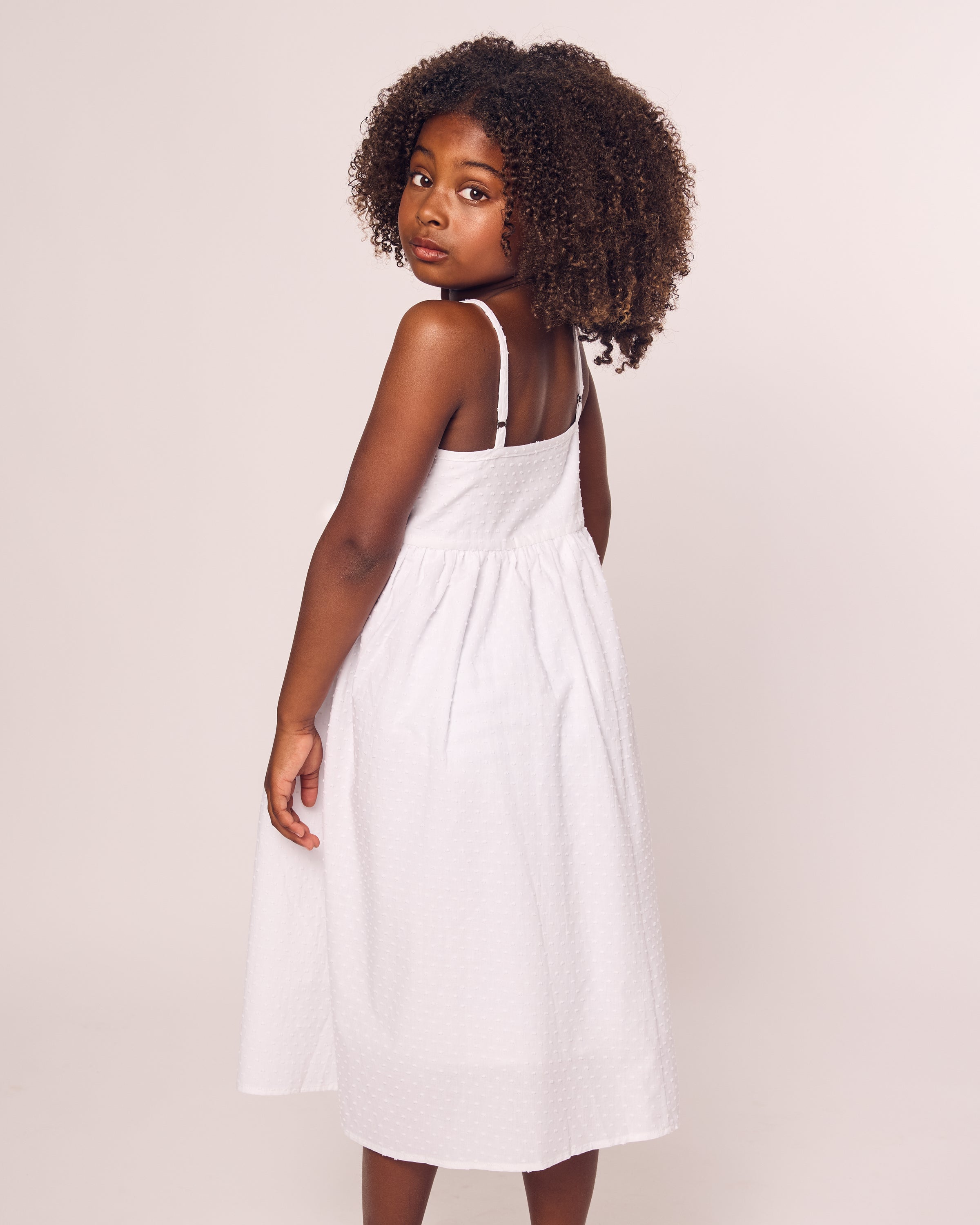 A young girl with curly hair wears the Petite Plume Girls Swiss Dots Serene Day Dress in White. She stands, looking over her shoulder at the camera, set against a plain off-white background that highlights the delicate texture of the cotton fabric.