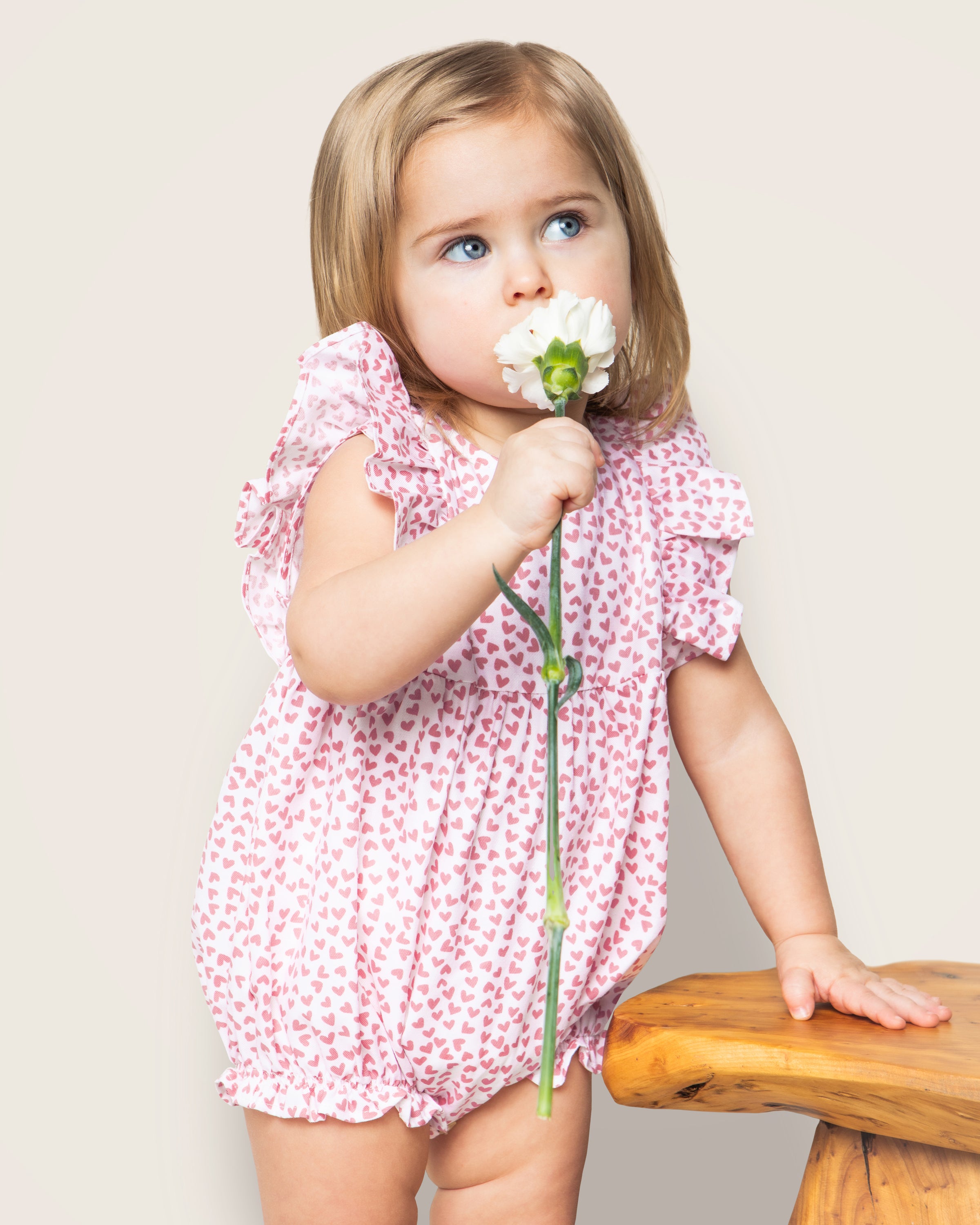 A small child with blonde hair wears Petite Plumes Babys Twill Ruffled Romper in Sweethearts. Holding a white flower, the child stands by a wooden surface, looking away from the camera.