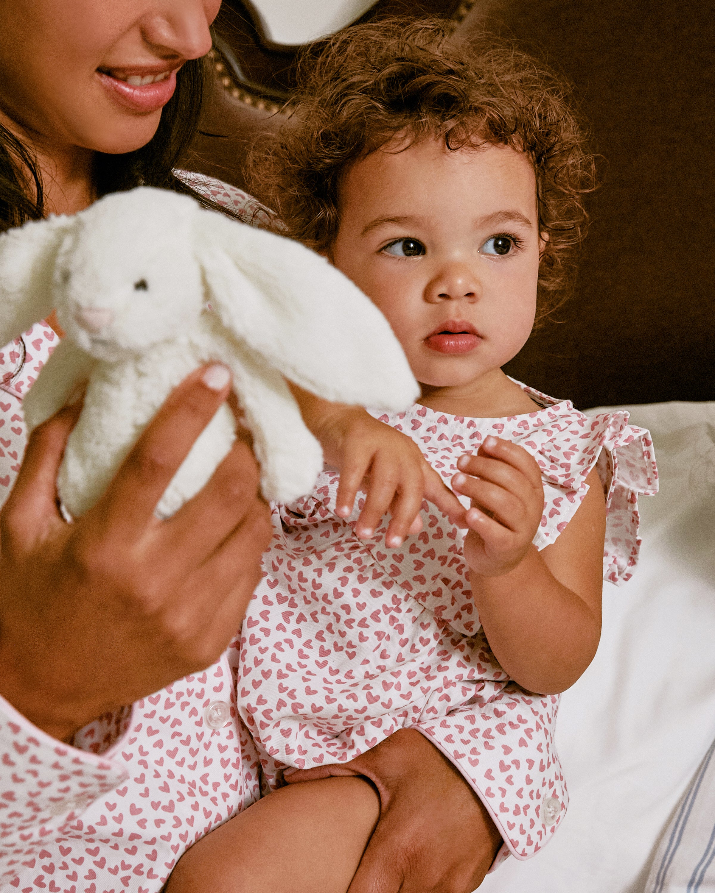 A woman and toddler sit together on a bed. The toddler, in Petite Plumes Babys Twill Ruffled Romper in Sweethearts, holds a plush bunny. The woman, also in heart-patterned attire, cradles the child securely on her lap.