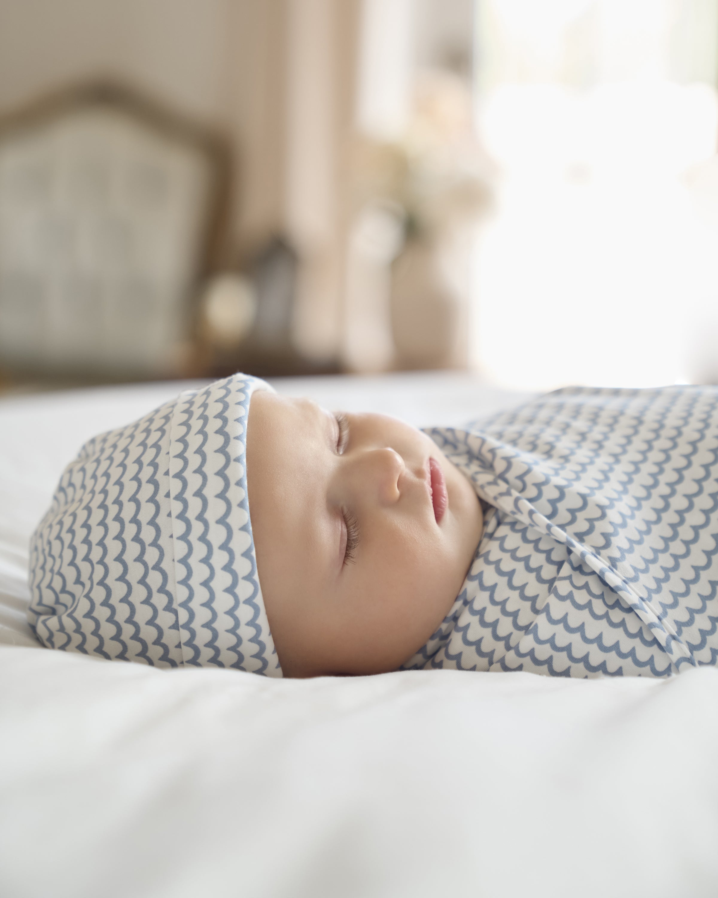 A baby sleeps peacefully on a bed, wrapped in Petite Plumes light blue and white patterned Pima Hat in La Mer, made from soft Peruvian Pima Cotton. The softly lit room creates a serene and cozy atmosphere.