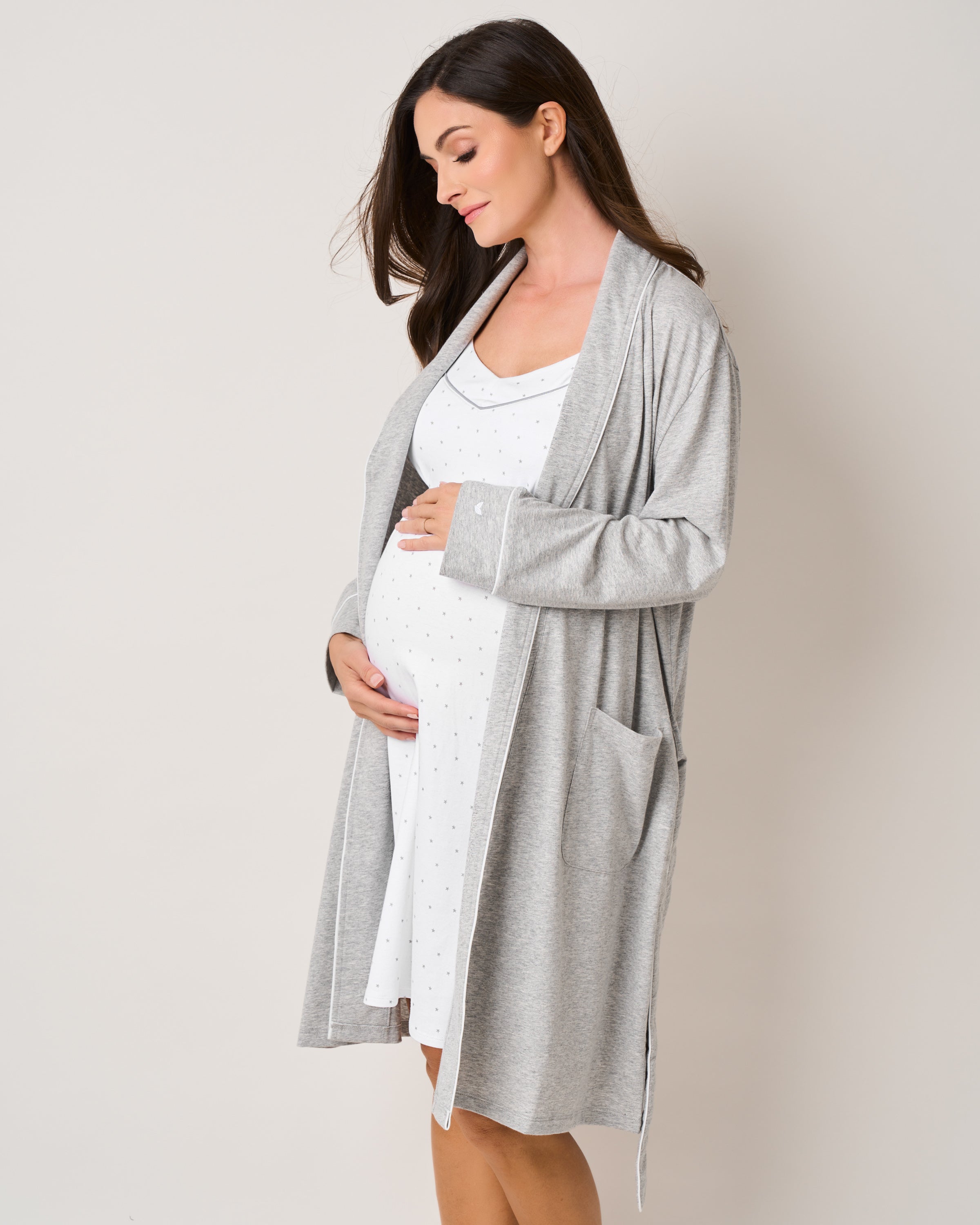 A pregnant woman in The Essential Maternity Set from Petite Plume in Light Heather Grey & Grey Stars gently cradles her belly against a plain backdrop. Her flowing brown hair enhances her serene expression, capturing the beauty of her pregnancy journey.