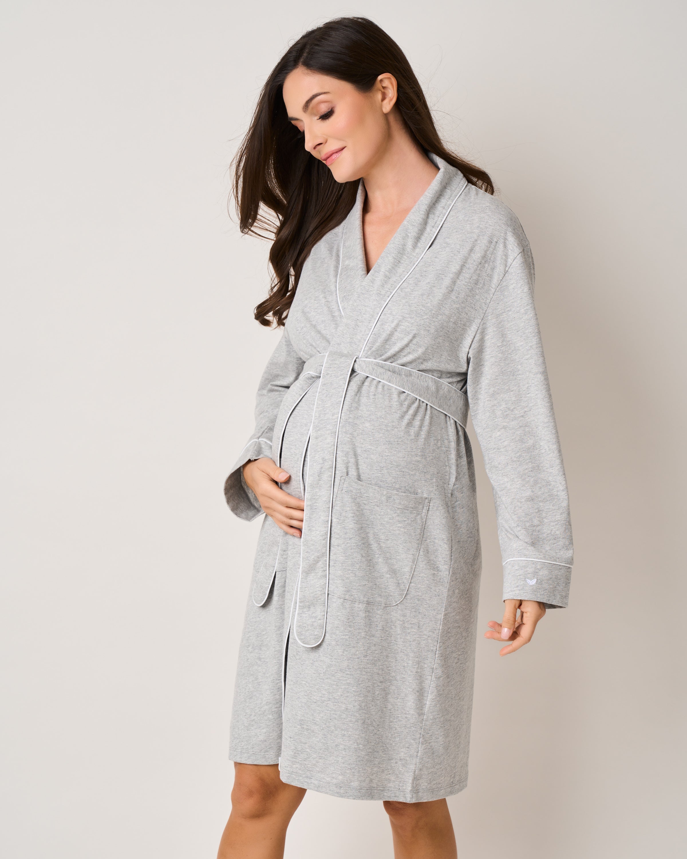 A pregnant woman embodies her journey, wearing Petite Plumes The Essential Maternity Set - Light Heather Grey & Grey Stars, with front pockets and a tied waist, gently cradling her belly. Her long hair drapes over her shoulders as she smiles softly against a plain, light-colored background.